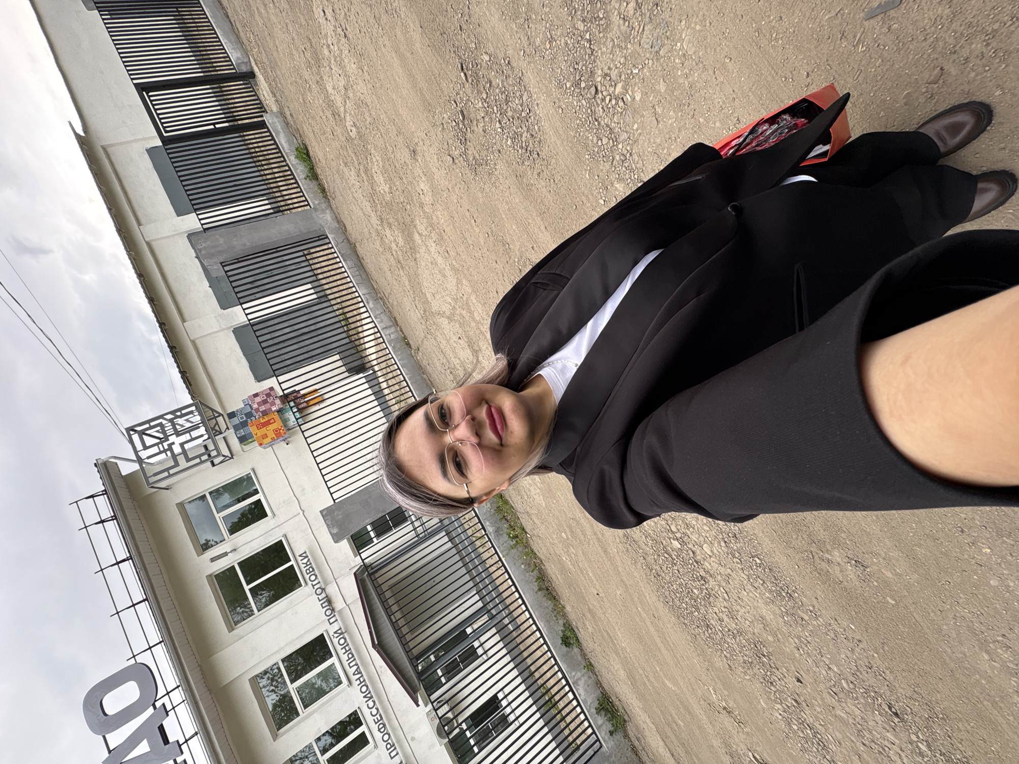 A woman in a black blazer, white shirt, and black pants stands outdoors on a gravel surface, likely a road or parking area. She is holding a red bag. She is wearing glasses and facing the camera, seemingly taking a selfie. Behind her is a light-colored building with multiple windows and a black metal fence running along the front of the building. A sign is visible on the building, with Russian text that indicates "ПРОФЕССИОНАЛЬНОЙ ПОДГОТОВКИ". The context suggests this could be a vocational training center or school. Based on the available text, the location is likely in Birobidzhan, Russia.