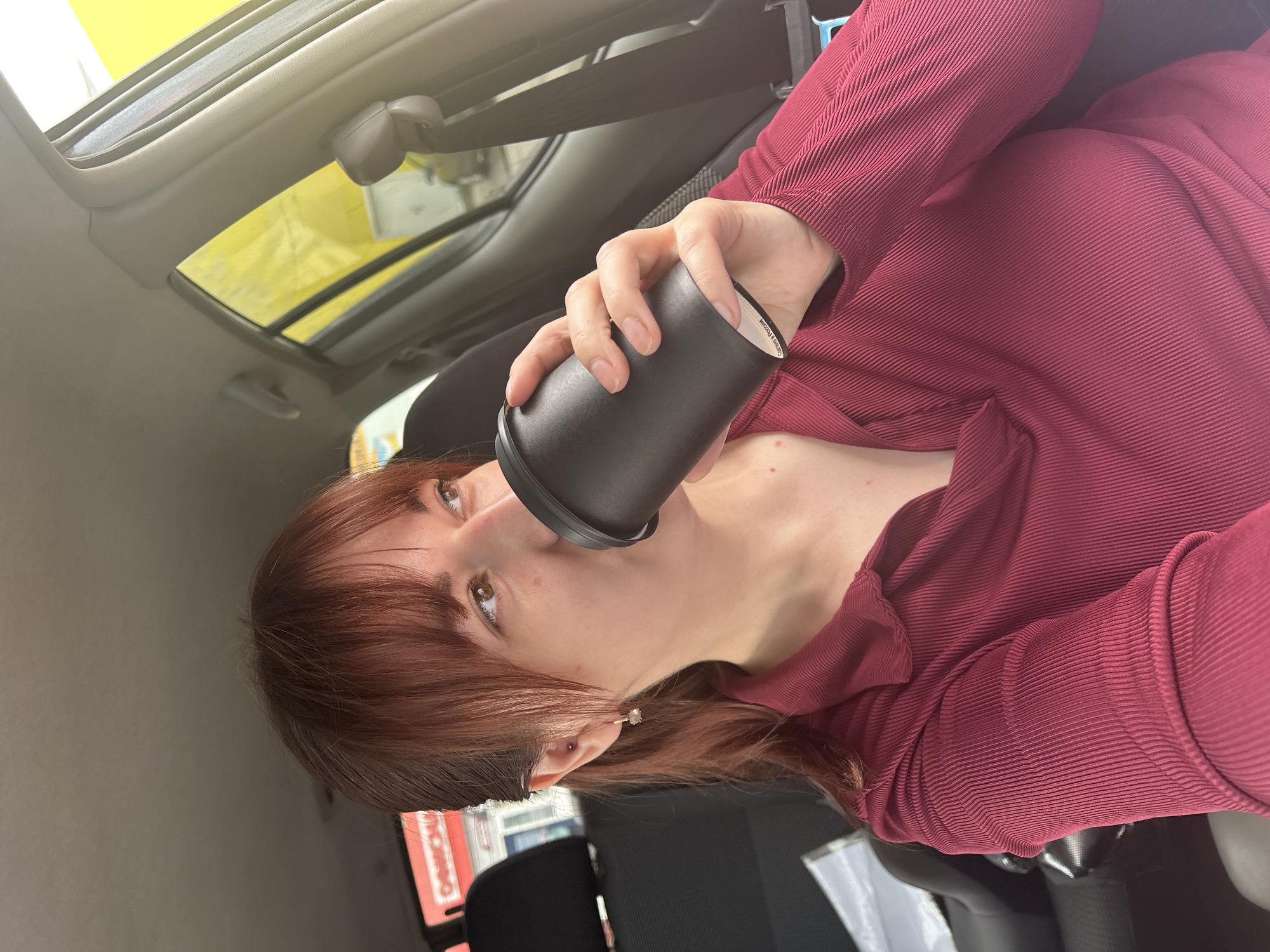 An adult female, estimated to be in her 20s or 30s, is depicted inside the driver's seat of a vehicle in Chaikovsky, Russia. She has light brown hair with bangs, pulled back into a ponytail, and brown eyes. The woman is wearing a long-sleeved, collared shirt in a deep red, ribbed fabric. Her left ear displays two piercings: a small stud on the helix and a small gold-toned hoop earring with a light-colored stone on the lobe. She is holding a cylindrical black reusable cup with both hands, positioned to her mouth as if drinking. The bottom edge of the cup features white text, visible as "Сделано в России" (Made in Russia).
The vehicle's interior features a beige headliner and A-pillar. A black headrest and part of a black seat are visible behind her. A dark grey seatbelt buckle and strap are visible on the right side of the frame. Through the rear window, a red sign with white Cyrillic text, partially legible as "ОПТИКА" (Optics), is visible above a building structure. Through the passenger side window, a large, bright yellow wall or structure is prominent, with a white building or object visible beneath it, and a faint outline of another vehicle in the distance.