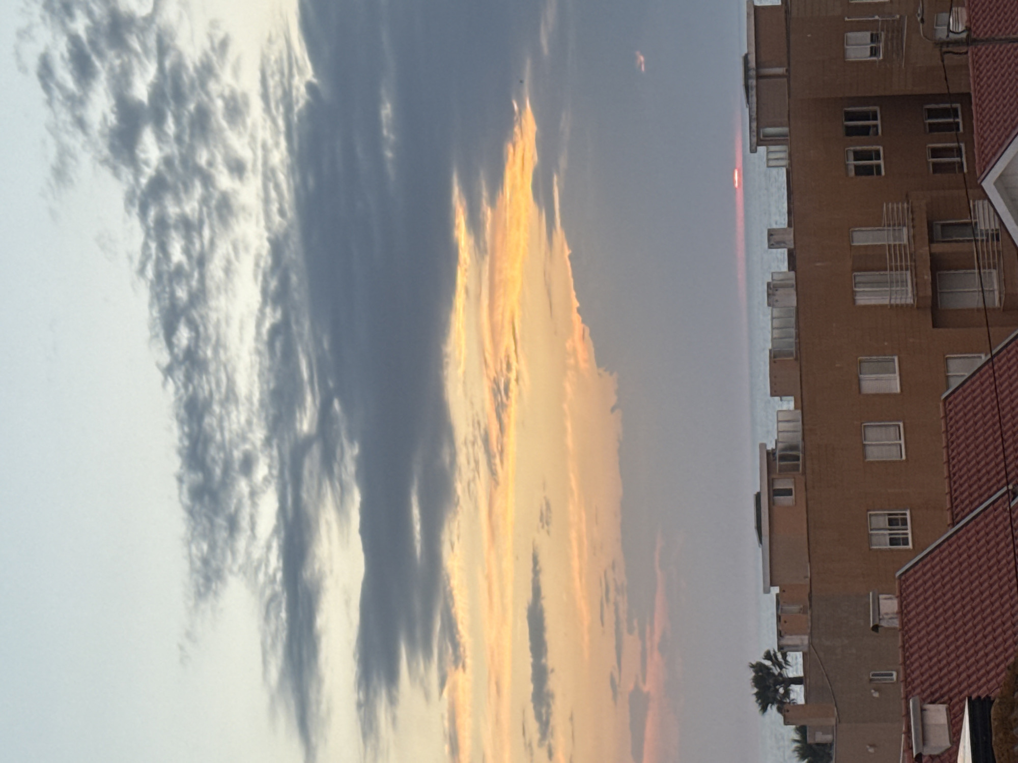 This media file captures a serene sunset over a coastal urban landscape. The sky dominates the left and upper portions of the image, displaying a rich array of colors and cloud formations. On the left, textured grey and white clouds stretch across the sky, while towards the center and right, the clouds are illuminated with warm golden and orange hues from the setting sun, creating a dramatic contrast.
A narrow, bright red-orange sliver, which is the setting sun, is visible just above the horizon line where the sky meets the calm, grey-blue sea. The sea appears as a horizontal band across the middle of the image.
To the right, several multi-story buildings line the coast. They are primarily constructed from a light brown or tan material and feature numerous windows, many with white frames, and some balconies. A building in the right foreground has a distinctive red-tiled roof. A few palm trees are visible near the base of the buildings, contributing to the coastal setting.
The image portrays the specific time of day as sunset, indicated by the vibrant colors in the sky and the visible sun disk. The weather appears to be partly cloudy. The location cues, including the sea, multi-story buildings, and palm trees, are consistent with a coastal urban environment like Vila Nova de Gaia, Portugal. The overall mood is one of tranquility and natural beauty. No visible text is present in the image.