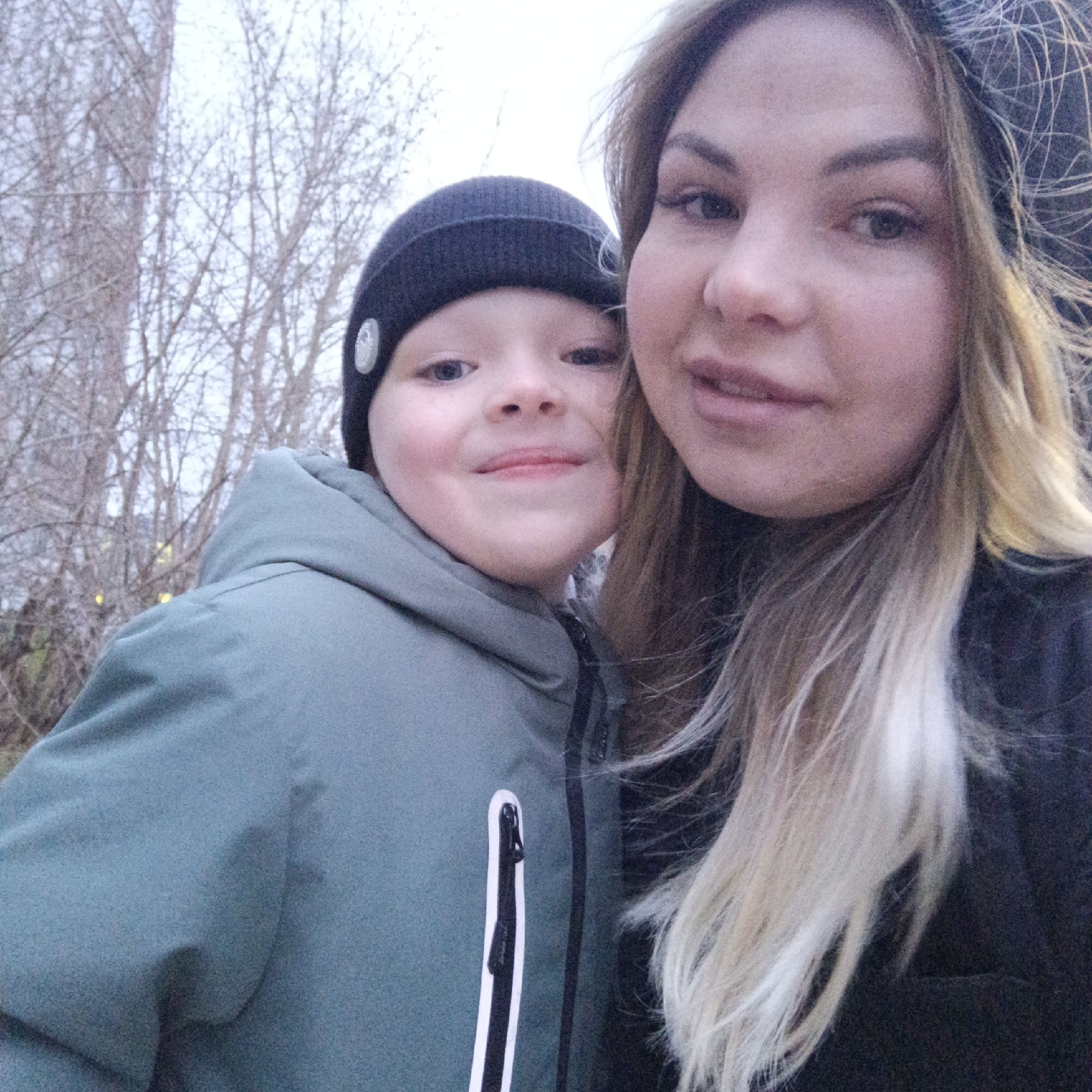 The image features two individuals, a woman and a young boy, captured in a close-up, likely selfie, outdoors. The woman, positioned on the right, has long blonde hair with darker roots and is wearing a dark beanie or hat. She has clear skin and appears to be wearing light makeup, with a subtle smile. The young boy, on the left, wears a dark knit beanie with a light-colored emblem or button on the side and a green or teal hooded jacket. The jacket has a black zipper with a distinct black and white zipper pull. He has light-colored eyes and a wider, joyful smile.
They are both looking directly towards the camera, posing for the picture. The background is filled with numerous bare tree branches against a light, possibly overcast sky. The absence of leaves on the trees, along with the warm clothing worn by both subjects (beanies and jackets), suggests a cold weather setting, likely late autumn or winter. The lighting appears soft and diffused, typical of an overcast day. No specific location landmarks are visible, only the natural setting of trees. There is no visible text in the image.