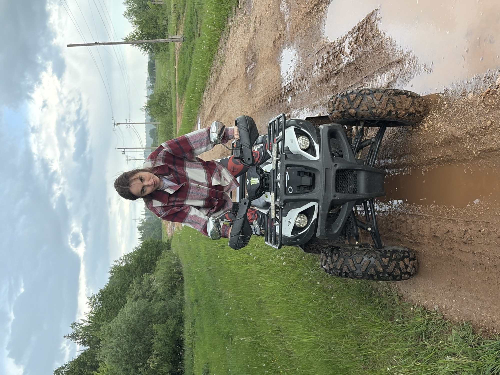 A female individual is on a black and white ATV. She is on a muddy dirt path. The landscape includes green grass, trees, and cloudy skies. Power lines are visible in the background. The location is in Nizhnii Vishur, Russia.