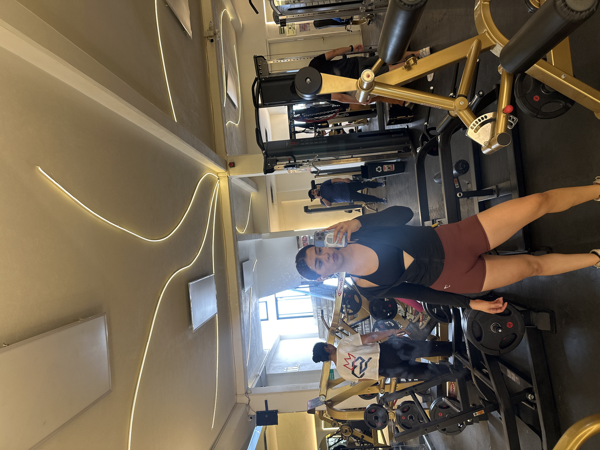 The image shows a mirrored view of a gym in Puebla, Mexico. The gym is equipped with weight machines. A woman in workout clothes holds a phone and takes a picture of herself. Another person is near her; also a man can be seen in the background exercising. The environment includes a mirrored wall with decorative lighting, and equipment.