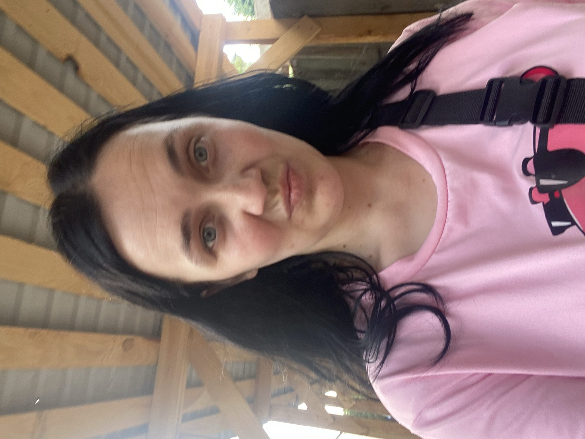 A medium shot from a slightly low angle shows a woman with long, dark hair and light blue eyes. She is wearing a pink t-shirt and a black strap across her chest. Her mouth is pursed, and she is looking directly at the camera. The background consists of wooden beams and a gray ceiling, suggesting she is under some sort of shelter, possibly outdoors. The lighting is natural and seems to be from above. The woman's skin appears slightly flushed.