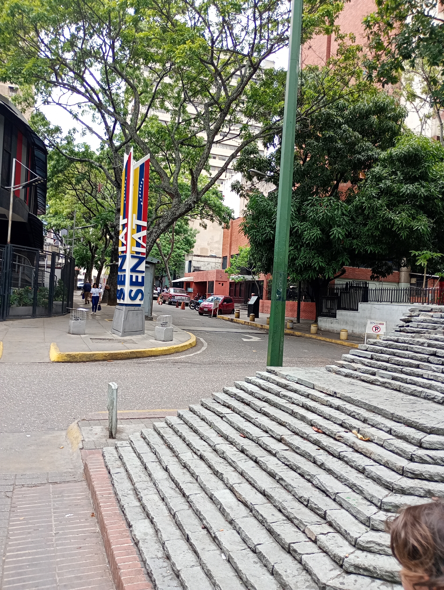 The image depicts a street scene in Caracas, Venezuela. The main subject is a large, vertical sign with the word "SENIAT" written on it, adjacent to a road. The colors of the Venezuelan flag are incorporated into the design of the sign.
People are walking on the sidewalk near the SENIAT sign. Vehicles, including cars and motorcycles, are parked or driving on the street. A set of wide stone stairs is visible on the right side of the frame. Trees line the street, and buildings are seen in the background. A green utility pole is prominently placed near the center of the image. The general environment suggests an urban setting.