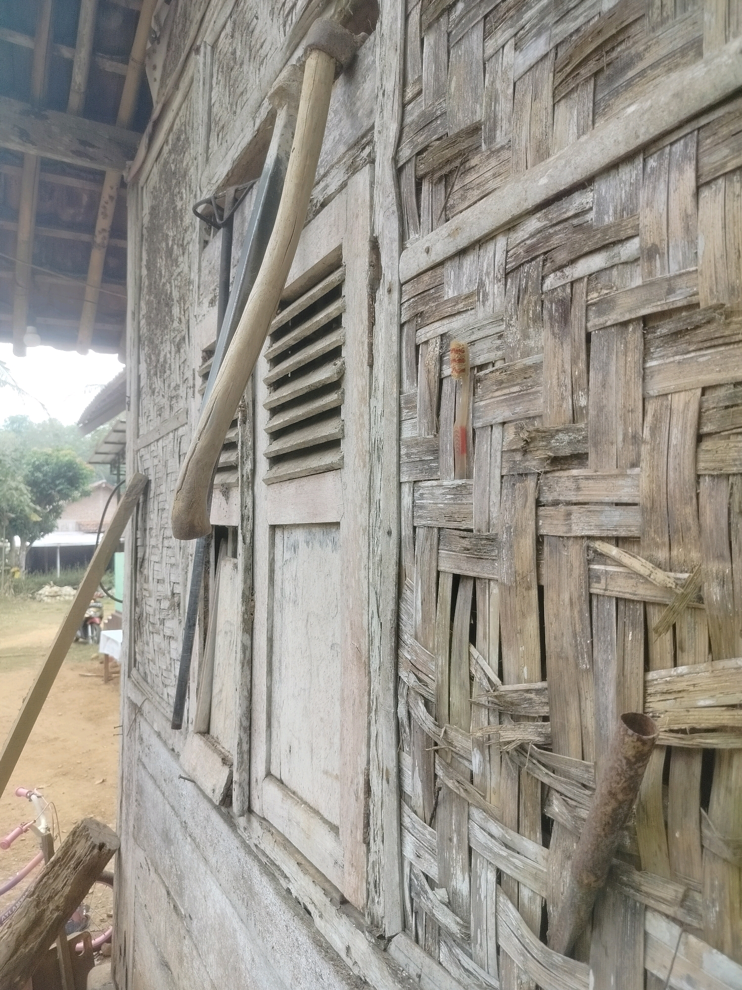 The image depicts the exterior wall of a traditional house in Pandansari, Indonesia, constructed from woven bamboo. A wooden hoe with a curved handle leans against the wall, its metal blade resting on a shuttered window. To the left, a longer wooden plank is propped against the structure, and further in the background, hints of a small dwelling and trees are visible. The woven bamboo wall shows signs of age and wear, with some pieces appearing loose or broken. A small, reddish-brown object, possibly a marker or decorative element, is embedded within the weave of the bamboo. To the right, a rusty metal pipe juts out from the wall. The ground in front of the house is dirt, and a portion of a pink bicycle is visible in the lower left corner. The overall scene suggests a rural setting.