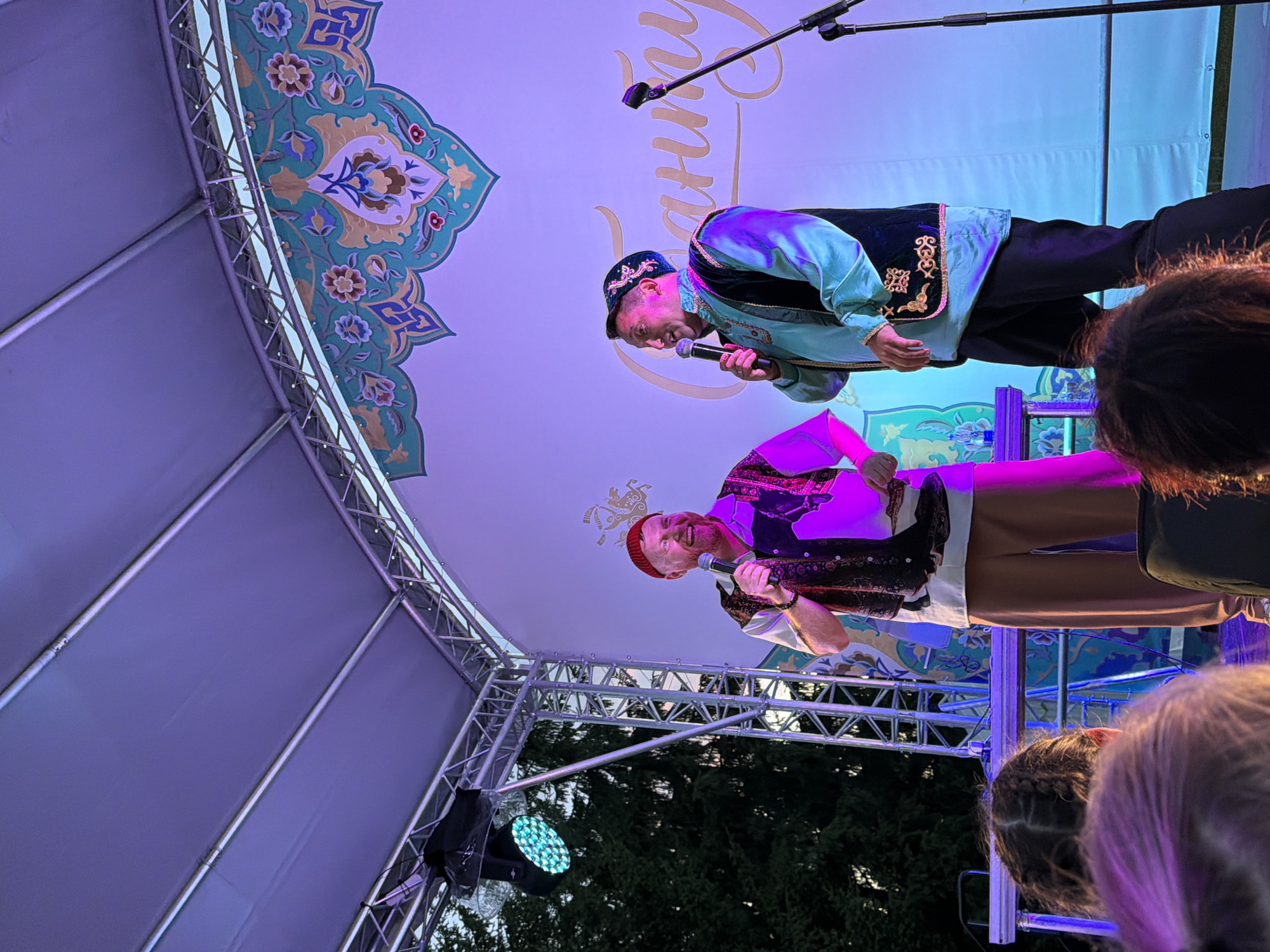 Two men are on a stage at an outdoor event. The man on the left, wearing a red hat and a patterned vest over a white shirt, holds a microphone and is smiling. The man on the right wears a dark cap with embroidery, a turquoise tunic, and a dark vest with gold trim. He also holds a microphone and is looking to his left. They are standing in front of a white backdrop with ornate green and blue decorations at the top and large, light-colored lettering in the center. A stage light is visible on the left, and trees and scaffolding are behind the stage. The foreground shows the backs of the heads of an audience, with purple and pink lighting illuminating their hair.