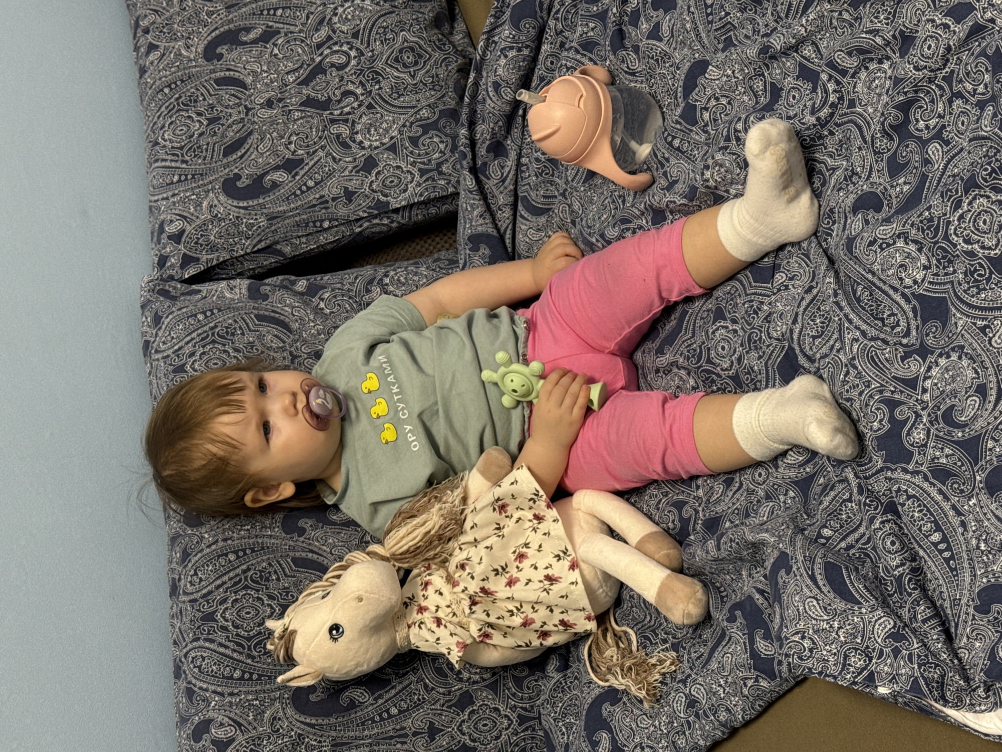 A toddler lies on a bed covered with a blue and white paisley patterned duvet. The child wears a light green t-shirt, pink pants, and white socks. They have a pacifier in their mouth and are holding a green toy. Next to the child is a stuffed horse wearing a floral patterned dress. A pink and clear sippy cup rests near the child's legs. The bed appears to be located indoors. There is no indication this picture was taken in Moscow, Russia.