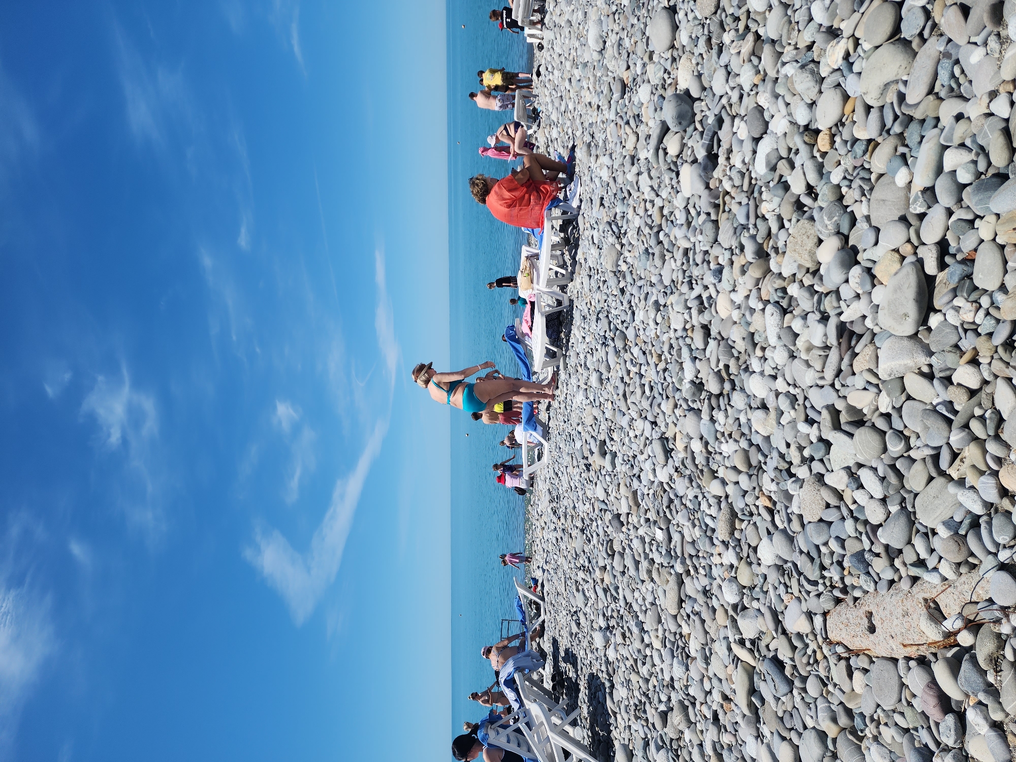 The image depicts a vibrant and sunny day on a pebble beach beside a tranquil blue sea. The sky is a clear, bright blue with scattered, wispy white clouds.
In the foreground, a vast expanse of smooth, grey and beige pebbles forms the beach. Among these pebbles, a weathered concrete-like object with some exposed rebar or wires is visible on the right side.
Several individuals are spread across the beach. In the mid-ground, a person wearing a blue two-piece swimsuit is standing, facing the sea, appearing to be either entering or exiting the water. Further along the shoreline, other beachgoers are visible; some are sitting on white beach chairs under what appear to be umbrellas, relaxing and enjoying the sun. One person is wrapped in an orange towel while seated. Others are standing closer to the water's edge. The people's attire suggests warm weather, with swimsuits and light clothing.
The sea stretches out calmly to the horizon, meeting the sky in a soft, unbroken line. The bright sunlight suggests it is midday or early afternoon. No visible text is present in the image. The scene conveys a relaxed and pleasant atmosphere typical of a summer day at the beach.