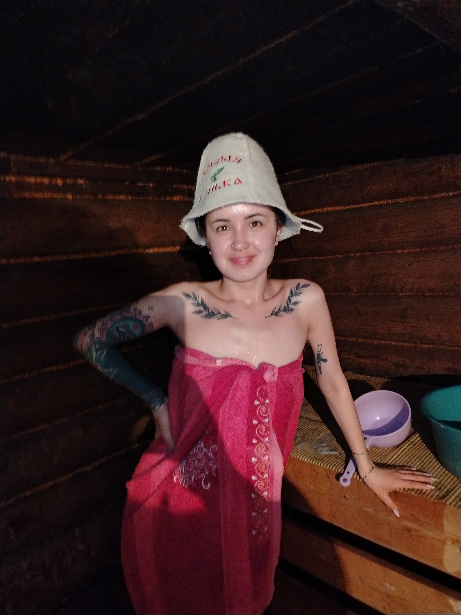 A single female individual is depicted within an indoor, wooden-paneled structure, consistent with a banya or sauna, located in Гожан, Russia. The subject, positioned centrally and facing the viewer, exhibits a slight smile. She wears a light-colored felt hat with red embroidered text reading "ДОБРАЯ БАНЬКА" and is wrapped in a pink towel featuring white embroidered patterns, covering her torso and lower body. Her skin appears moist. Visible tattoos include a full sleeve on her left arm with multi-colored designs, two branch-like motifs on her clavicles, and a stylized firearm on her right upper arm. A septum piercing is present. Her left hand is placed on her hip, while her right hand, adorned with a beaded bracelet, rests on a light-colored wooden bench to her right. On this bench, a woven mat, a purple plastic bowl, a teal plastic bucket, and a purple plastic ladle are present. The environment is dimly lit with warm tones, reflecting off the dark, horizontally paneled wooden walls.