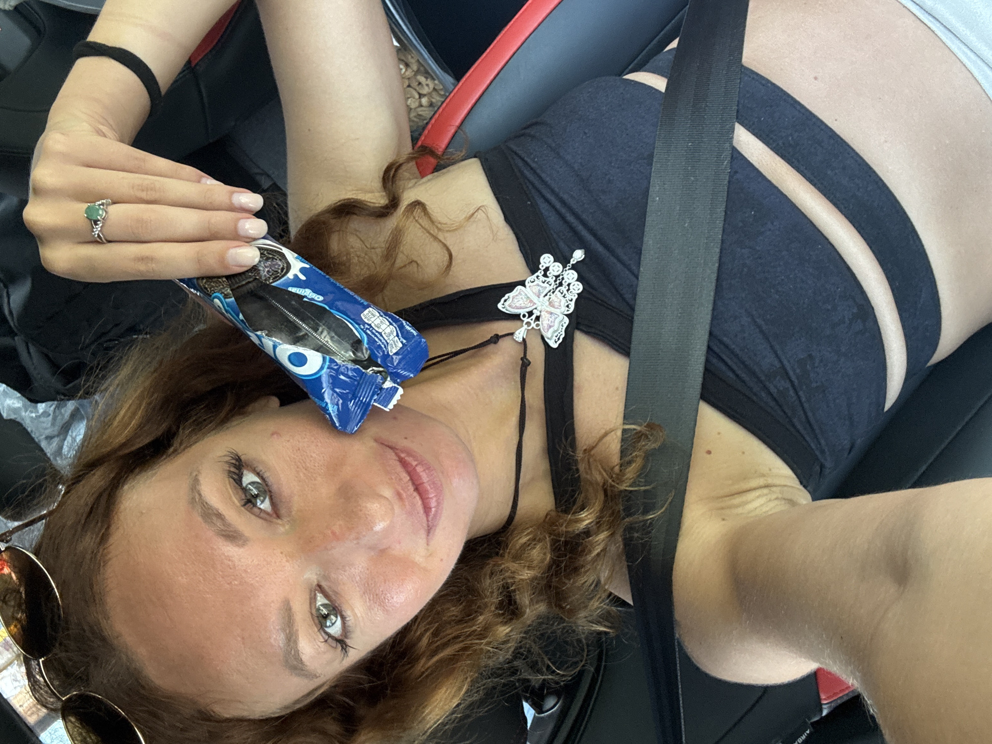 A woman is lying in a car, secured with a seatbelt, and holding a package of Oreo cookies. She is wearing a black top and has a butterfly brooch pinned to the fabric near her neck. Sunglasses rest next to her on the left. A ring adorns a finger on her left hand. The setting is inside a car, indicated by the seat and seatbelt. The location of Lũng Phìn Commune, Vietnam, is not evident in the image.