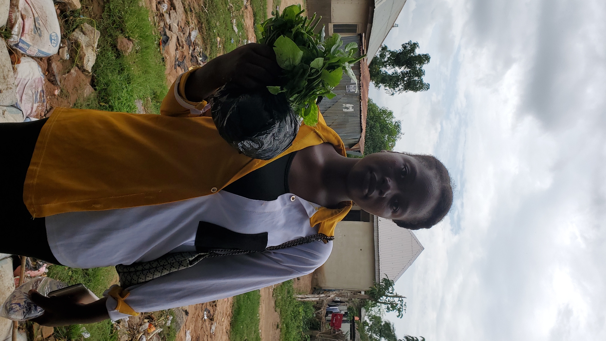 A young woman stands outdoors in Jalingo, Nigeria, holding a bunch of leafy green vegetables wrapped in black plastic in her left arm. She is wearing a color-blocked shirt, with the left side being yellow and the right side white, and a dark-colored shirt underneath. A patterned shoulder bag hangs from her right shoulder. In her right hand, she holds a clear plastic bottle of water. The background shows a few buildings with corrugated metal roofs, some trees, and a cloudy sky. The ground is covered in dirt, rocks, and scattered debris.