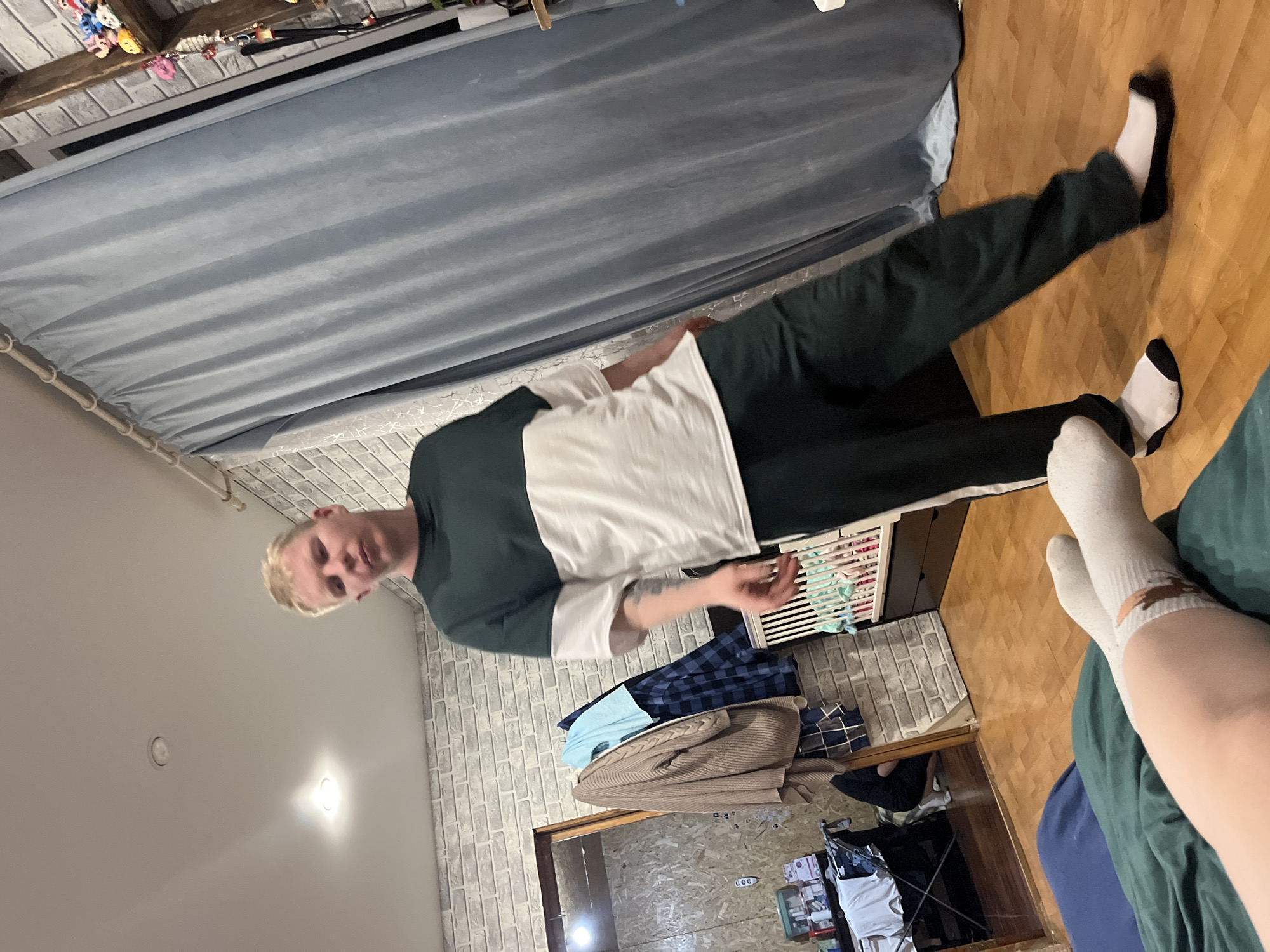 The image shows a person standing in a room. The person is wearing a two-toned green and white shirt, dark pants, and socks. One leg is raised. A bed with a gray curtain is behind the person. The floor is wooden. In the background, there is a brick wall and a glimpse into another room with clothes and boxes. The location appears to be a room with personal belongings, possibly a bedroom. The room's design suggests an interior setting, but the image does not provide specific details to identify a location in Vesyoloye, Russia. There are no identifiable landmarks, street signs, or other location markers in the image.