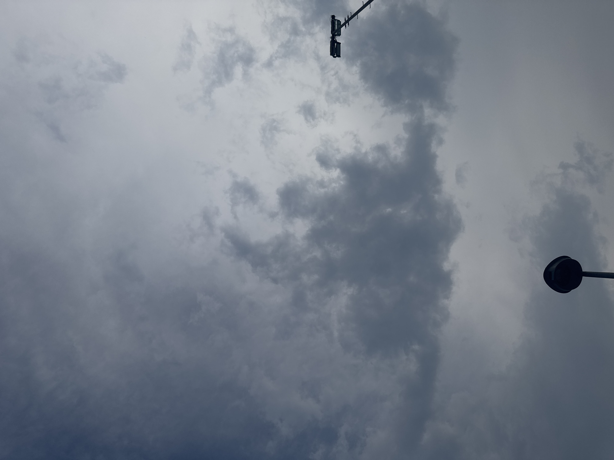 The image shows a cloudy sky over Chiasso, Switzerland. In the lower left portion of the frame, a dark, bulbous streetlight is visible, mounted on a pole. In the upper right, a tall pole supporting multiple floodlights is present, angled upwards. The sky is filled with a variety of clouds, ranging from dark, heavy formations to lighter, more diffused ones, suggesting overcast conditions. No people are present in the image.