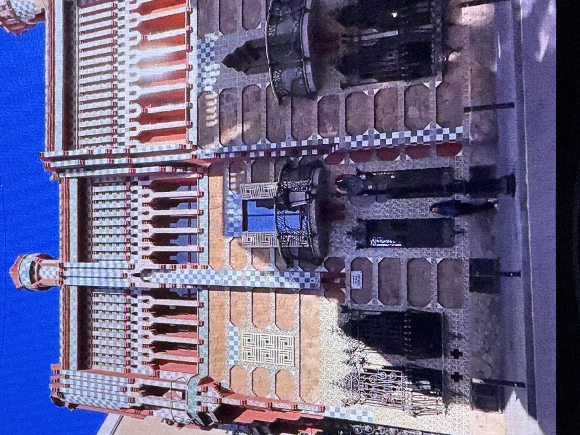 This image displays an overhead view of a building with intricate architectural details, situated in Dubai, United Arab Emirates. The building features a facade with a repeating pattern of red and white checkered tiles, interspersed with sections of warm-toned material and decorative grilles. Several balconies or recessed areas are visible along the structure, some with ornate metalwork. A prominent cylindrical tower with a checkered pattern extends upwards on the left side of the building. The overall design appears to be a blend of traditional geometric patterns and modern construction.
There are no people visible in the image. The scene suggests a clear day, given the strong shadows cast by the building, indicating it is likely daytime. The architecture, particularly the detailed tilework and the overall design, is a notable aspect of the scene. The location cue "Dubai, United Arab Emirates" further contextualizes the image, suggesting a modern urban setting. No visible text is present in the image.