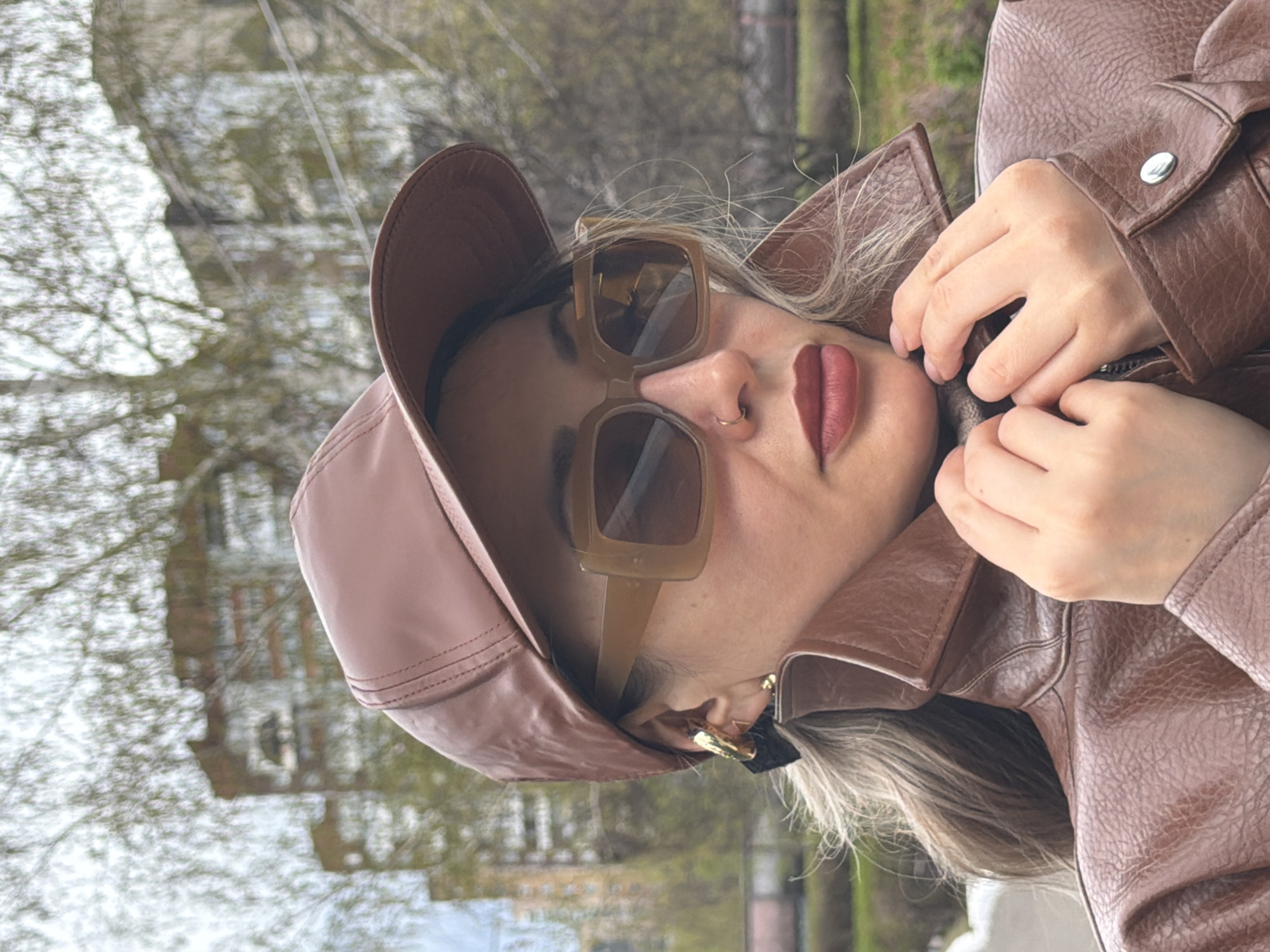 A woman wearing a brown leather jacket and matching hat and sunglasses is seen outdoors. She has a nose ring and lipstick. Her hands are near the collar of the jacket. Trees and a building are visible in the background, which suggests an urban environment. It is not possible to confirm this is Krasnoyarsk, Russia with the given information.
