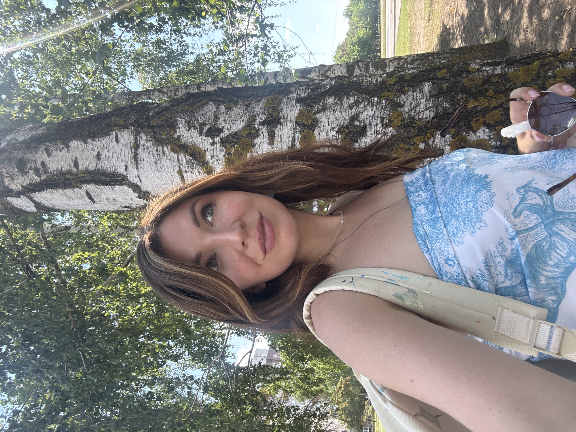 A young woman with long, brown hair is taking a selfie outdoors, leaning against a birch tree. She is wearing a white dress with a blue toile-like pattern and a light-colored strap across her shoulder, likely part of a bag. She is holding a pair of sunglasses in her right hand, which is near her face. The woman has a pleasant expression, with a slight smile.
The scene is set in a natural environment, with lush green trees and foliage visible. The sun is shining, casting a warm glow and creating some lens flare. The bark of the birch tree is white with dark markings and patches of lichen. In the background, beyond the trees, hints of buildings and a road suggest an urban or suburban setting. The image appears to have been taken during the daytime, likely on a sunny day. The overall mood is relaxed and cheerful. No visible text is present in the image.
