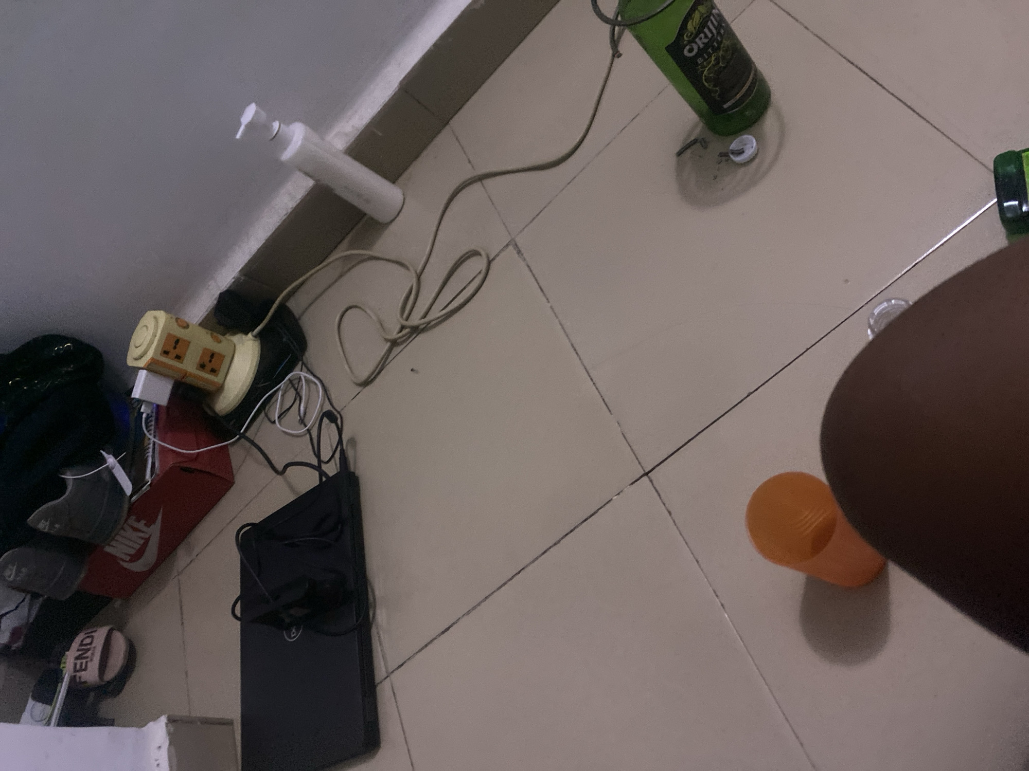 The image depicts a floor in what appears to be a domestic setting in Howayi, Nigeria. The floor is tiled with light-colored square tiles, some of which show signs of wear and tear. Various objects are scattered on the floor, suggesting a state of disarray or recent activity.
Prominently visible are several electronic items and accessories, including a power strip with multiple sockets, several chargers, and cables. A black laptop is also present, alongside a red Nike shoe box. Other items include a white bottle with a dispenser top, an orange cup, and the bottom of a green bottle, possibly indicating that drinks were consumed. A portion of a person's leg and arm are visible on the right side of the image, suggesting someone is present but not the primary focus.
The lighting suggests it is daytime, though the overall illumination is dim, possibly due to indoor lighting or overcast conditions. There are no strong indicators of the specific time of day or weather outside. The presence of personal belongings like shoes and a laptop points to a living space. The visible text includes "NIKE" on the shoe box and "FENDI" on what appears to be a shoe or accessory at the bottom left. The brand name "ORIGIN BITTER" is partially visible on the green bottle. The scene conveys a sense of everyday life, with a hint of clutter.