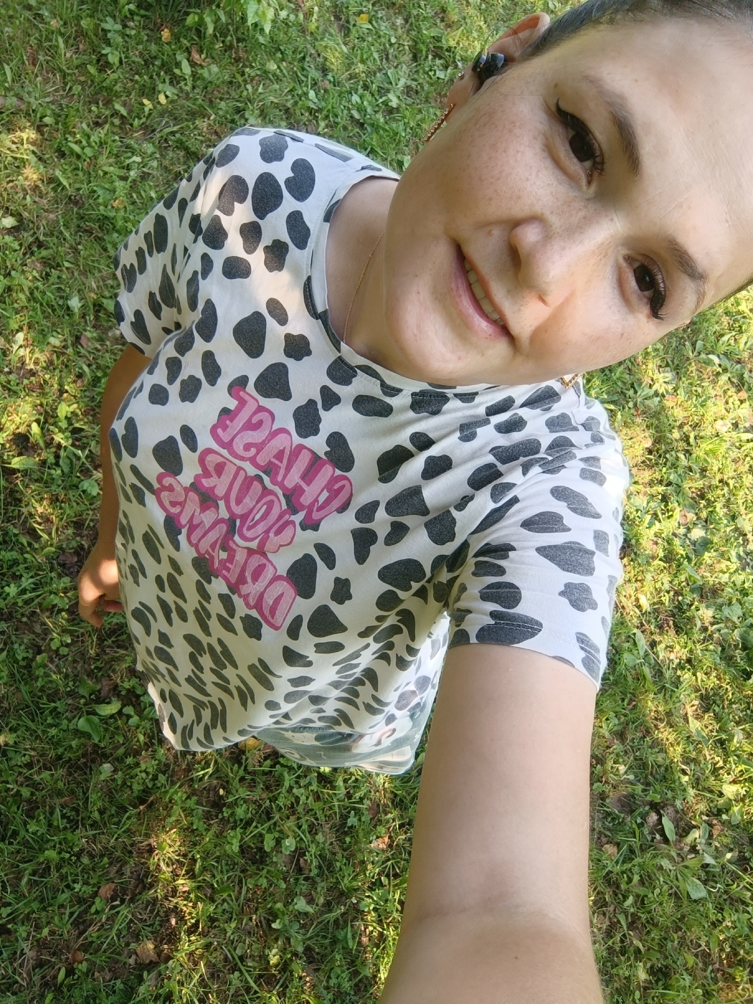 A high-angle, medium shot captures a woman with light skin and dark hair from the chest up, posing for a selfie outdoors. She is wearing a white t-shirt with a black and gray dalmatian-like pattern and the words "CHASE YOUR DREAMS" printed in pink lettering. Her face is tilted towards the camera, and she is smiling. She has dark eyeliner and light brown eyes, and freckles are visible on her cheeks. She is wearing black earbuds in her ears. The background consists of green grass, with dappled sunlight creating shadows and highlights. The image quality is somewhat grainy, and the lighting is bright. The location is an outdoor grassy area.