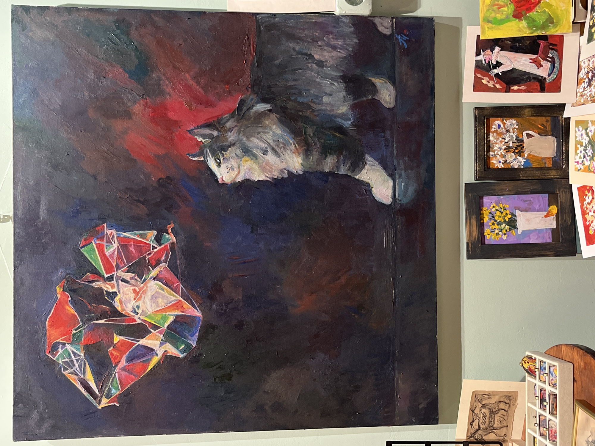 This is a photograph of an oil painting displayed in a retail setting. The painting, which is the primary subject, depicts a fluffy gray and white cat standing on a dark, reflective surface. The cat is looking upwards and to the left at a large, abstract, geometric shape composed of red, yellow, green, and purple facets. The background of the painting is dark and textured, with washes of red and blue. The painting appears to be signed in the bottom right corner with red paint.
Below the main painting, several other smaller artworks are visible, including framed paintings of flowers and a drawing of a goat. A white tray containing several small, colorful rectangular objects, possibly charms or buttons, is also on display. The overall impression is of an art gallery or a market stall exhibiting original artwork.
