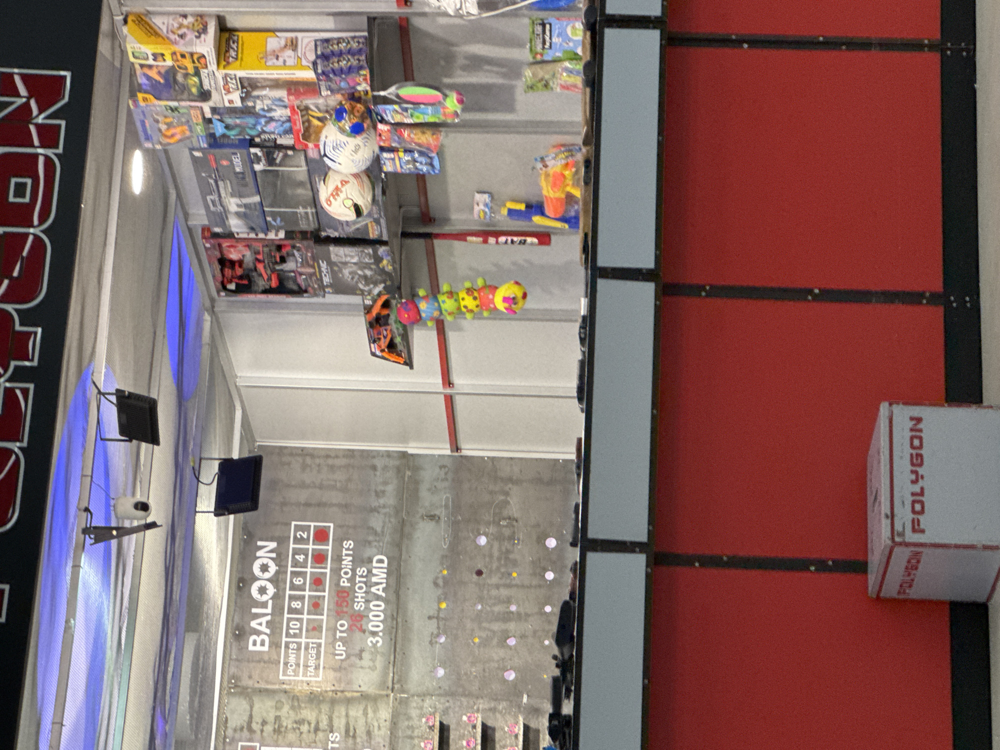 This image captures an indoor game booth, likely within an arcade or entertainment center in Yerevan, Armenia. The scene is brightly lit by artificial lights, including some blue accent lighting.
On the left side of the booth, various prizes are displayed on shelves, including boxed toys such as Lego Technic model kits, other scale models, two white soccer balls, a colorful caterpillar plush toy, a red baseball bat, and a yellow and blue water gun.
The central feature is a game wall marked with "BALOON" (likely "BALLOON"), detailing scoring "POINTS 10 8 6 4 2" for different "TARGET" sections. The game offers "UP TO 150 POINTS" for "26 SHOTS" and costs "3.000 AMD," indicating the currency of Armenia. The grey target wall features several purple and yellow circular targets. Above the game wall, small monitors and what appears to be a camera or sensor are mounted.
To the right, red wall panels are visible, and a white box with the word "POLYGON" printed in red is placed on a ledge. A large, partially visible sign on the far left also displays the "POLYGON" brand name. The overall setting suggests an engaging, prize-winning activity at an entertainment venue.