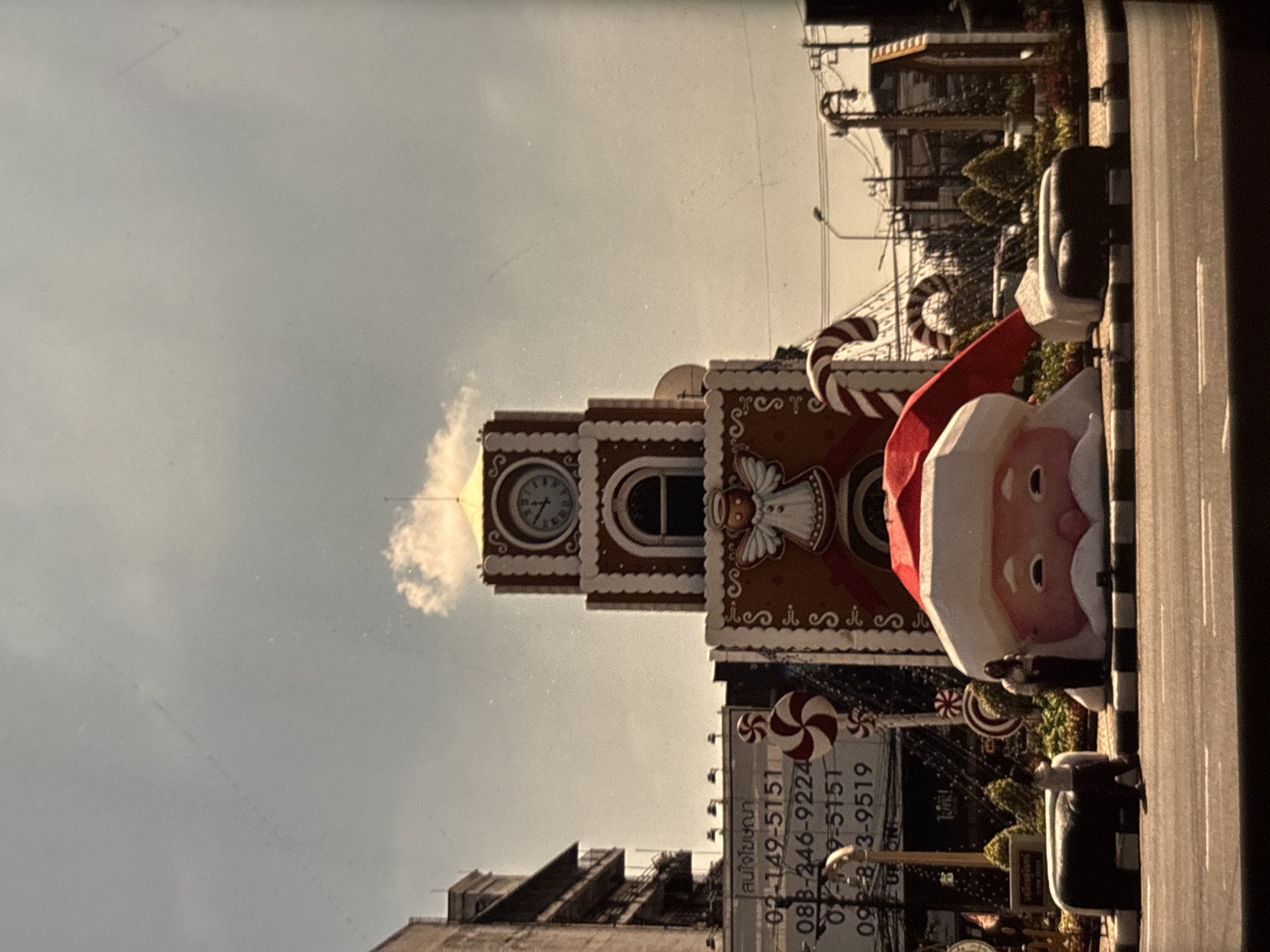 In Chalong, Thailand, a gingerbread-themed clock tower stands as a prominent Christmas decoration. The tower features a clock, candy cane designs, and an angelic figure. In front of the tower, a large inflatable Santa Claus head with a red hat and white trim is positioned on the street. A woman stands near the Santa head, and another person is visible near a black bench-like structure. The scene is set against a backdrop of buildings, streetlights, and utility poles under a cloudy sky. The street in the foreground shows pedestrian crossings and vehicle traffic is implied by the presence of the road.