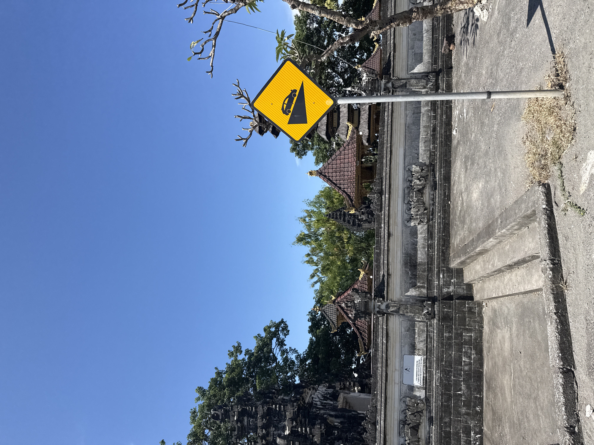 A vertical, daytime shot shows a road sign near a Balinese stone structure, possibly a temple.

The sign is yellow with a black diagram of a car driving down a steep hill. The sign is mounted on a metal pole on a concrete sidewalk.

Behind the sign, there is a carved Balinese stone wall with intricate designs, partially obscured by trees. The wall has decorative roof structures, possibly part of a temple or gate.

The sky is bright blue, suggesting clear weather.

The location is in or near Batununggal, Indonesia, based on the presence of Balinese architecture.