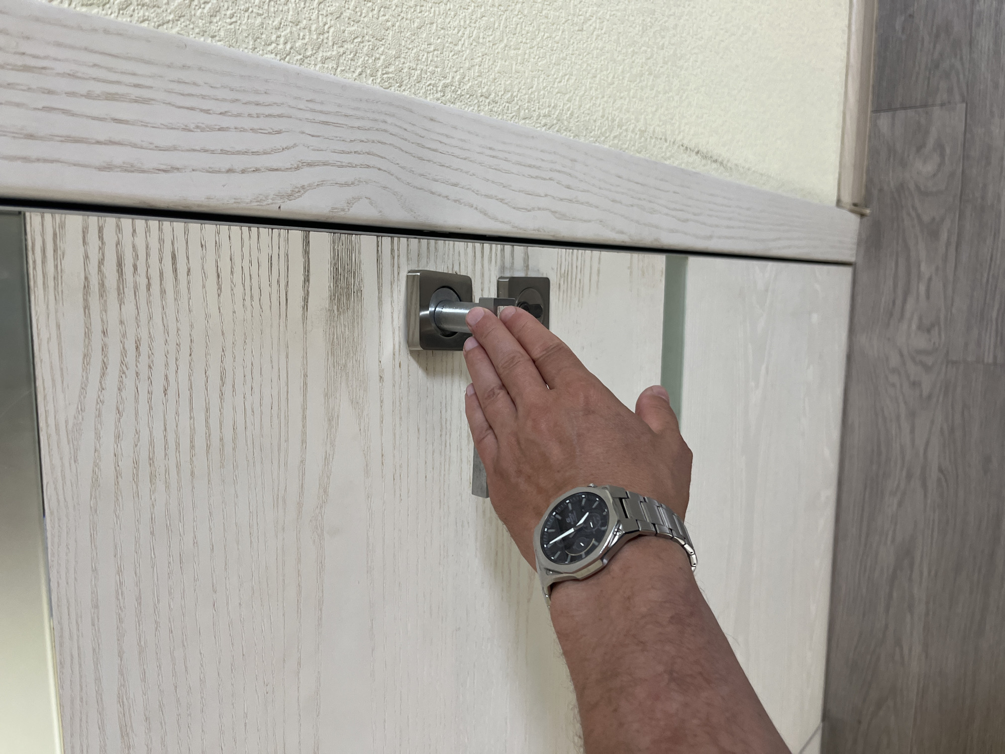 A person's left hand, wearing a silver-banded wristwatch with a black face, is shown pressing down on a modern, silver-colored door handle. The door itself has a light, wood-grain pattern, and the handle is attached to a white door. The door frame, also with a light wood-grain pattern, is visible on the right side of the image, adjacent to a textured, light-colored wall. The floor below is covered in grey wood-grain laminate flooring. The image appears to be taken indoors, possibly in a residential or commercial building.