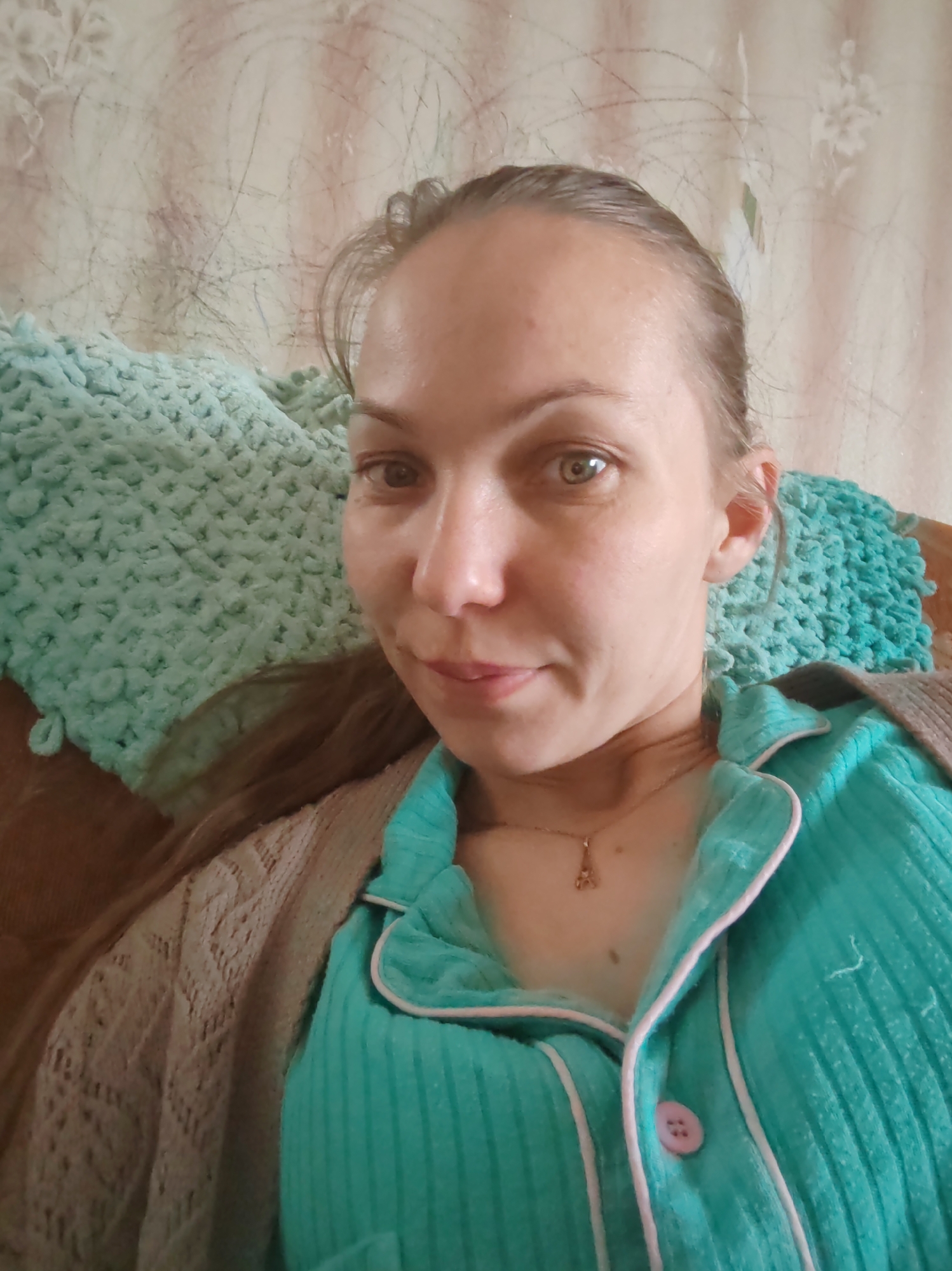 A medium close-up selfie captures a woman reclining in an indoor setting, likely a home in Чухлэм, Russia. She has light skin, light-colored eyes (appearing greenish-brown), and light brown hair tied back or pulled away from her face. Her expression is calm and neutral, with a subtle, slight smile.
She is wearing a casual, ribbed long-sleeved top in a turquoise or mint green color, featuring white piping along the placket and a single visible light pink button. Over her shoulders, she has draped a light brown or beige knitted cardigan with an intricate openwork pattern. A delicate gold-toned necklace with a small Eiffel Tower pendant hangs around her neck.
A plush, textured blanket or throw in a matching turquoise or mint green color, possibly crocheted or knitted, partially covers her. Behind her, a light-colored wall is visible, adorned with a faint, faded pattern and numerous dark, thin, scribble-like marks, possibly from pencil or crayon. The overall scene conveys a relaxed and comfortable atmosphere. No text is visible in the image.