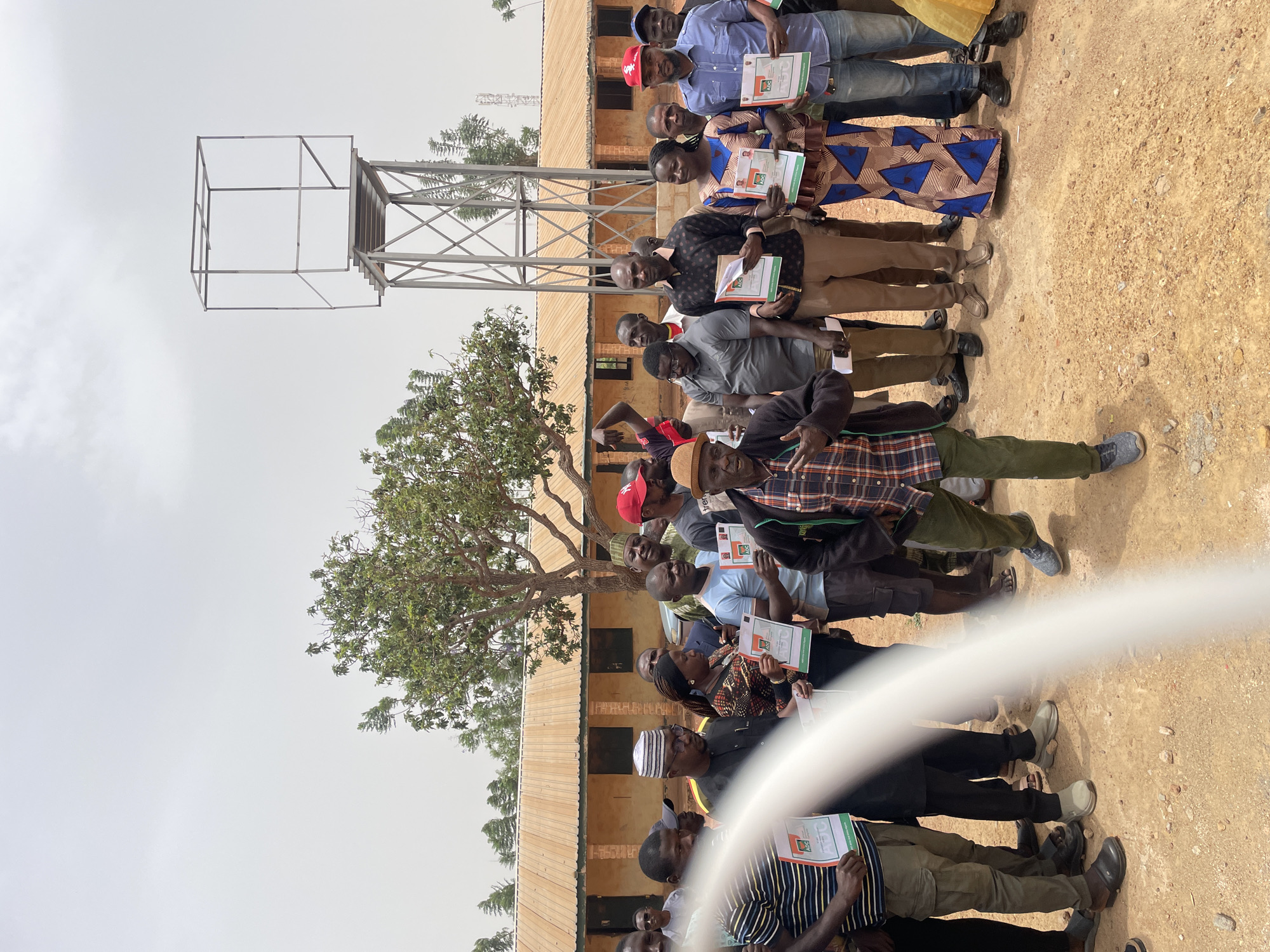 A group of approximately 20 individuals stand in a row outdoors on a sandy, dirt ground. Each person holds a document, possibly a certificate, with green and red colors visible on it. The people are dressed in a variety of casual clothing.
Behind the group, there is a single-story building with a brown roof and light-colored walls. A metal water tower structure stands to the left of the building, and a tree with green leaves is visible in front of it. The sky is overcast.
A blurred white object, likely a hose or rope, obscures the lower center of the image.
The location may be in Jos, Nigeria, due to visual cues related to the architecture and landscape.