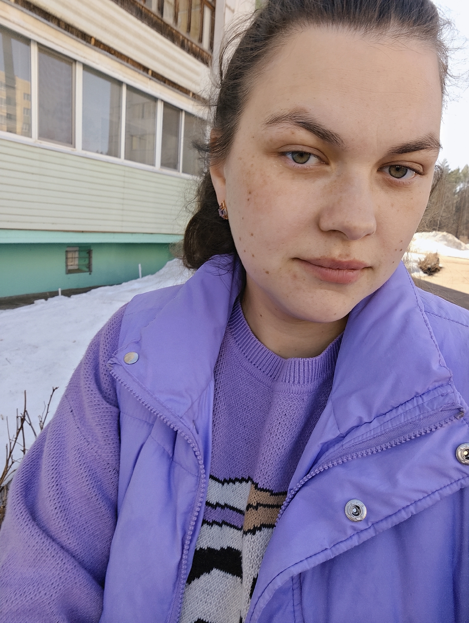 A person, presumably female, is captured in a selfie. The individual is facing forward with fair skin, brown hair pulled back, and brown eyes. She has freckles on her face and is wearing a purple sweater and a matching purple puffy vest. Her top appears to have some beige, black, and white coloring. She is also wearing earrings.
The setting appears to be outdoors in a location where there is still snow on the ground. Behind the subject is a building with multiple windows. A small patch of snow is visible on the ground. Given the prompt, the image was most likely taken in Chaikovsky, Russia.