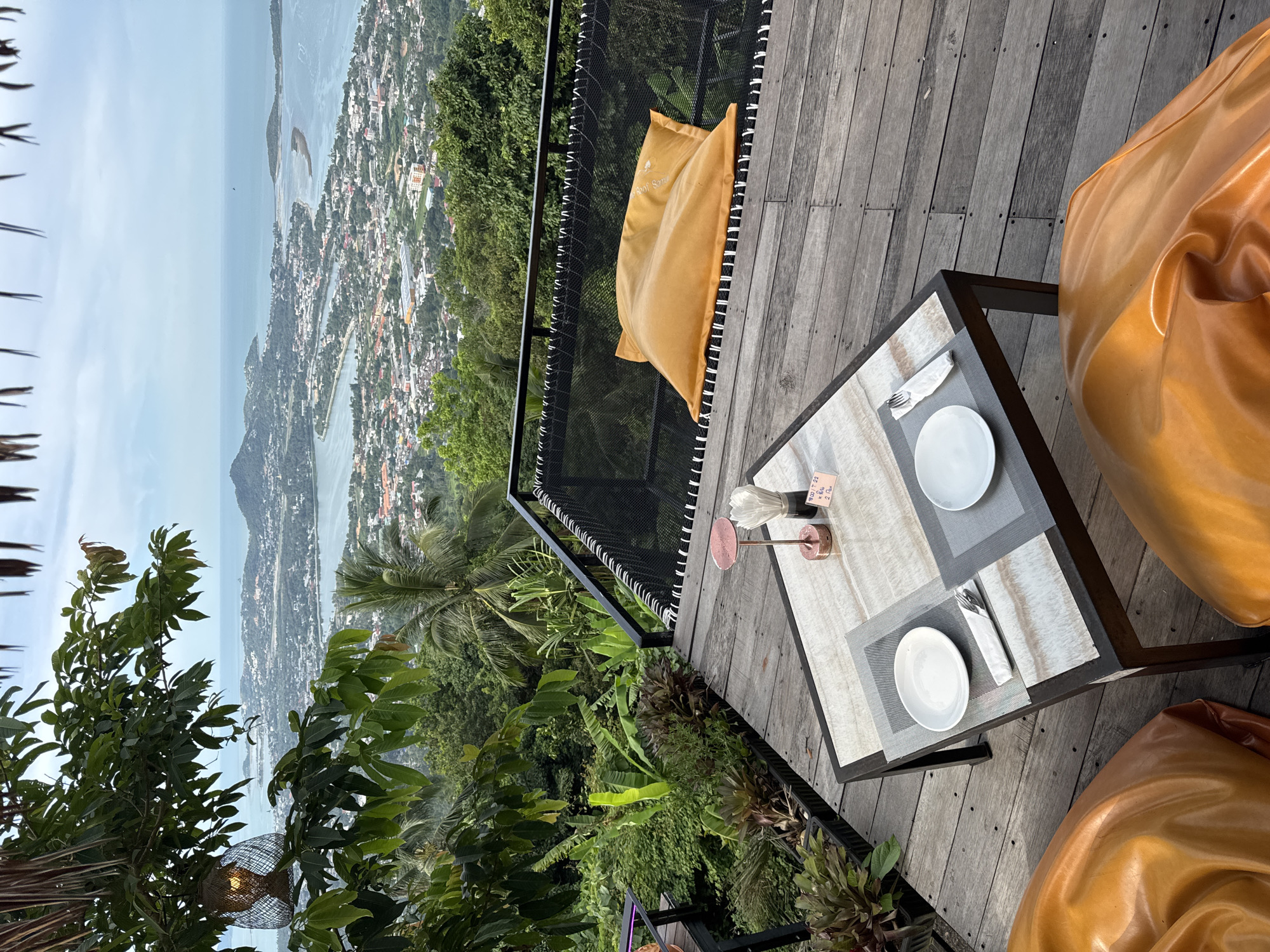An outdoor dining area with a scenic view of a coastal city and ocean. In the foreground, a wooden deck features a small table set for two, with white plates, cutlery, and a napkin. Beside the table, two large, orange beanbag chairs are visible. Beyond the deck, a black netting hammock holds an orange cushion. Lush green foliage and palm trees surround the area, creating a tropical atmosphere. In the background, a sprawling city with buildings of various colors sits on hillsides overlooking a calm, blue ocean with a distant island. The sky is overcast. A woven lantern hangs from a tree branch on the left side of the frame.
