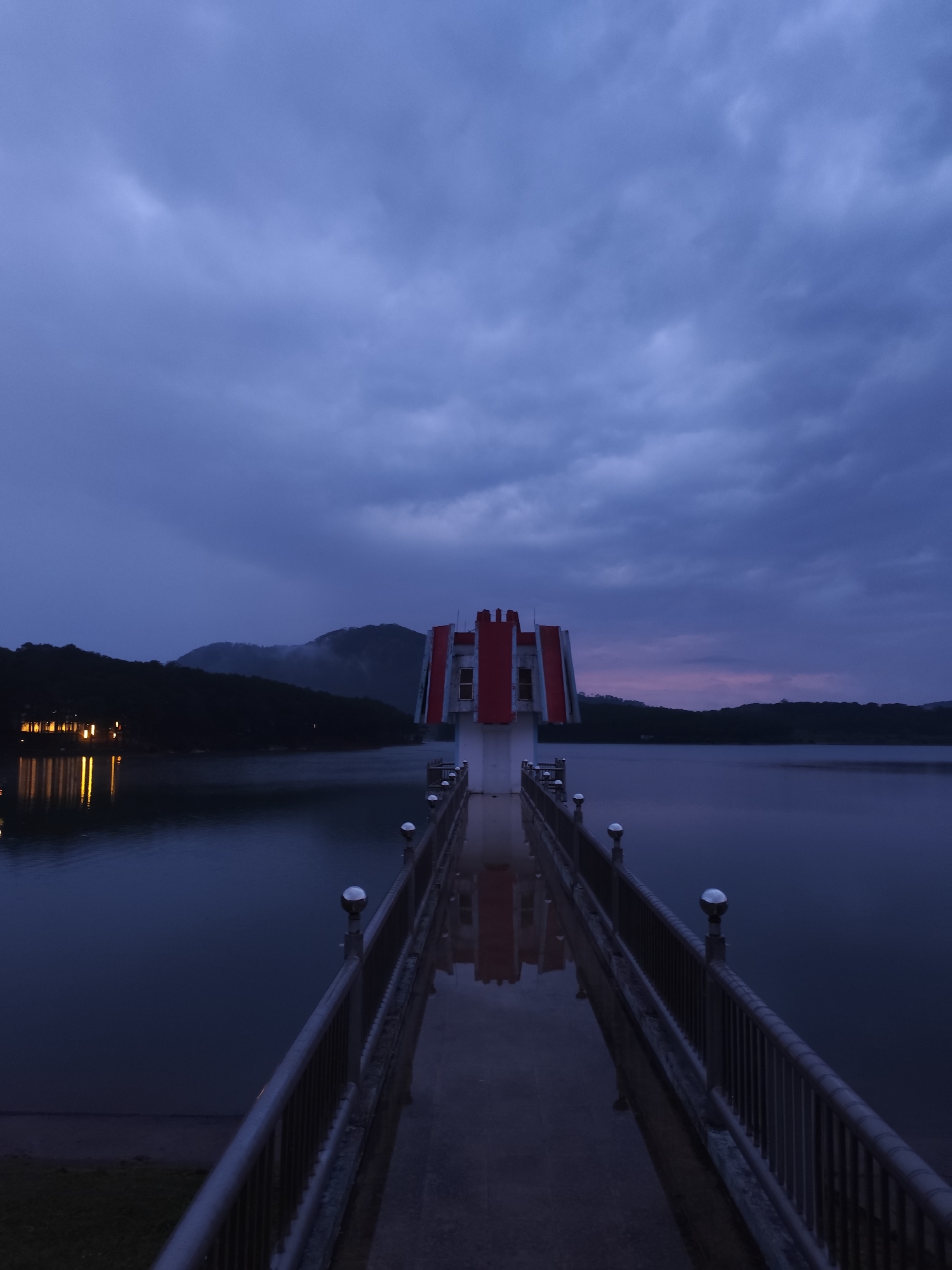 The image presents a serene twilight or dawn scene over a body of water, likely a lake or reservoir, in Da Lat, Vietnam. A long, concrete pier extends from the foreground into the distance, leading to a distinctive structure with white walls and striking red elements, possibly roofs or decorative panels. This structure is centrally positioned and perfectly reflected on the wet surface of the pier.
The pier features dark metal railings with reflective spherical finials lining both sides, creating a strong leading line towards the building. On the left side of the scene, distant artificial lights from a settlement or buildings are visible on the far shore, casting warm yellow reflections onto the calm water. Dark, forested mountains or hills form a silhouette against the horizon in the background.
The sky is heavily overcast with dense, textured clouds in shades of dark blue and purple, indicating either late dusk or early dawn. A subtle reddish-purple glow is discernible on the horizon to the right, hinting at the fading or emerging light. The overall lighting is low, creating a quiet and contemplative atmosphere. There are no people visible, and no specific activity is taking place, contributing to the tranquil stillness of the moment. No discernible text is visible within the image.