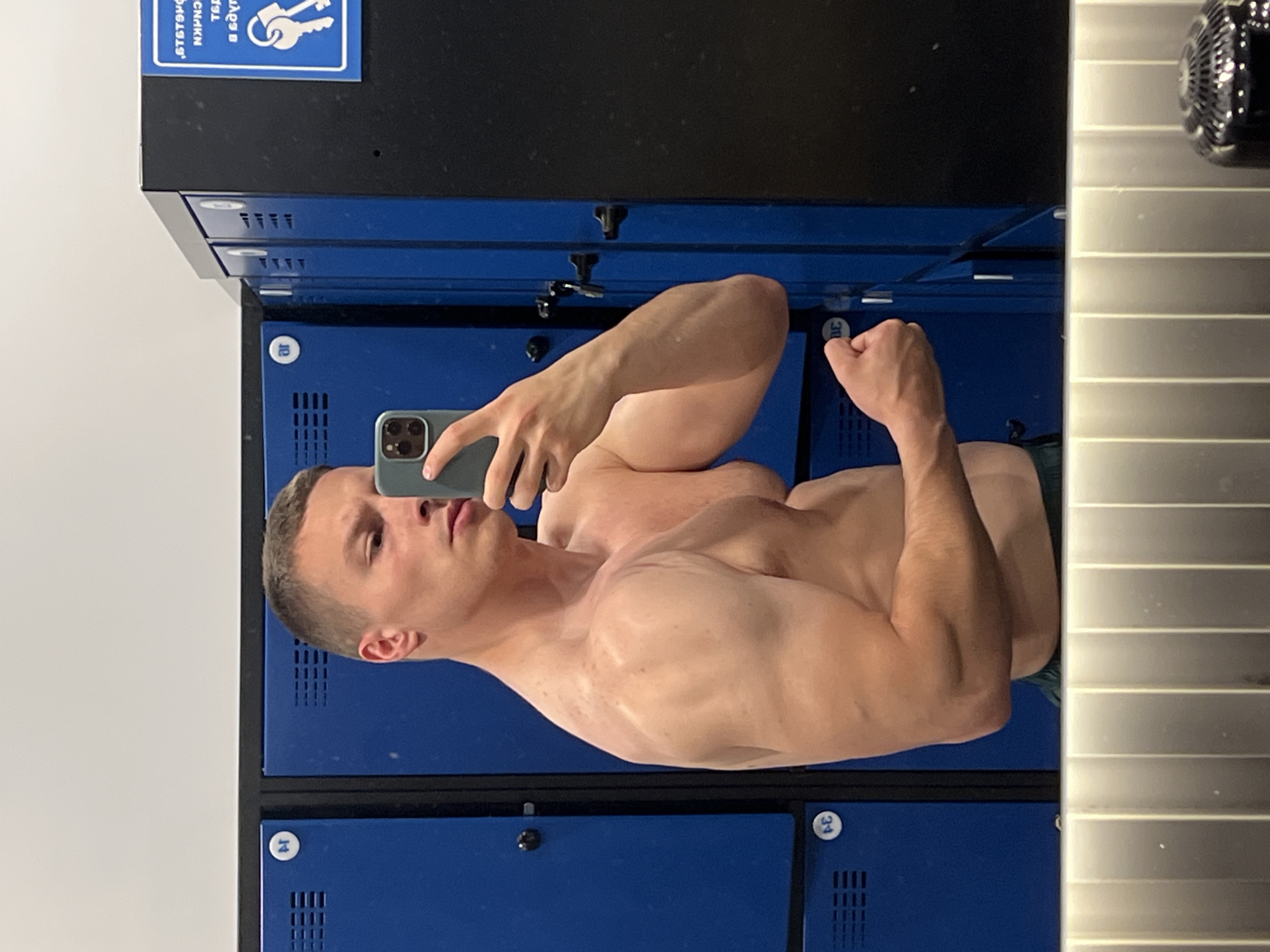 A shirtless man takes a selfie in front of a locker room mirror. The lockers are blue with silver numbers and are located in Burgas, Bulgaria. The man is holding a smartphone with a multi-lens camera. His right arm is flexed.