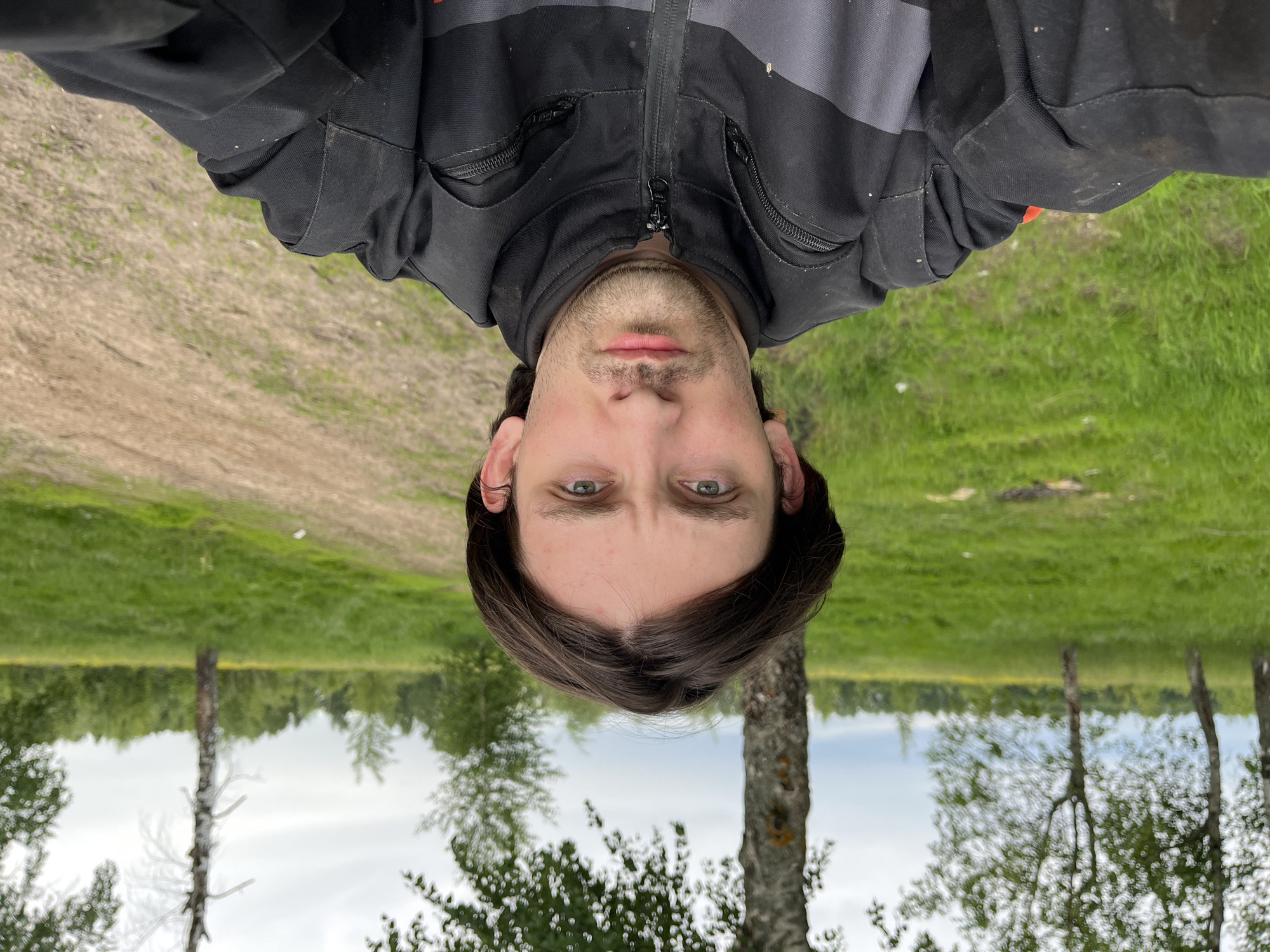 A male person is upside down in the foreground. He has short, dark hair and green eyes. He is wearing a black and gray jacket. Behind him is a landscape featuring grass, trees, and a body of water reflecting the sky. The location is Новочувашский, Russia.