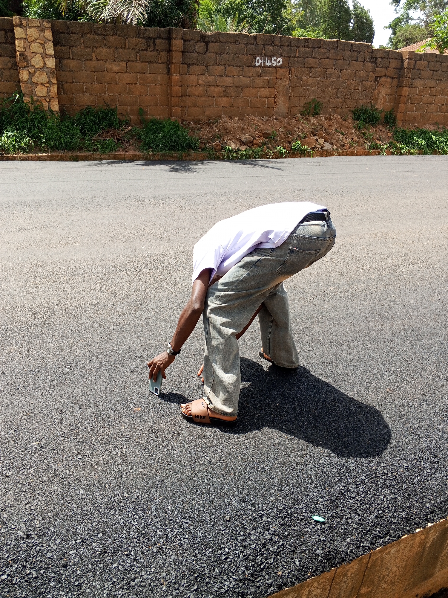 In Bukuru, Nigeria, a person is bent over on a paved road, holding a smartphone. The person is wearing a white t-shirt, grey jeans, and brown slide sandals. Their body is angled downwards, and their right hand is holding the phone near the ground. The road surface is dark asphalt with small pebbles. In the background, a brick wall runs horizontally, with some vegetation and trees visible above it. A number, "01450", is painted in white on the wall. The scene suggests a casual moment or activity taking place outdoors on a street.