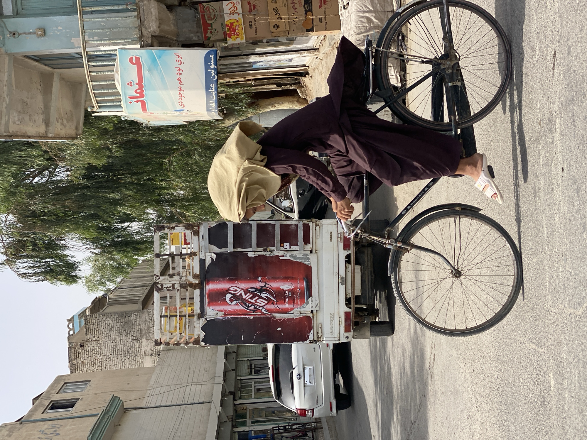 A young individual, wearing a dark brown traditional shalwar kameez and a light brown headscarf, rides a bicycle down a paved street in Kandahar, Afghanistan. The individual's feet are clad in white sandals. The bicycle has a black frame, a rear rack, and spoked wheels. The individual is leaning forward, appearing to pedal.
To the left of the cyclist, a white SUV is parked. Behind the SUV, a small truck with a white utility body is also parked. The truck's rear is decorated with a large, faded advertisement for "Sting" energy drink, featuring a red can design. The truck's license plate is not clearly visible.
Buildings line the street on both sides. On the right, a two-story building features a blue and white sign with Arabic script and a balcony with a railing. Stacks of packaged goods, possibly snacks, are visible in front of the building. On the left, a building with a light-colored facade has several windows and a white rolling shutter on the ground floor. Overhead, power lines are strung across the street. Trees with dense green foliage are visible, one prominently to the left of the blue and white sign. The sky is overcast with a hazy, bright atmosphere. The street surface is asphalt with visible cracks and debris. The overall scene depicts an everyday street view in an urban environment.