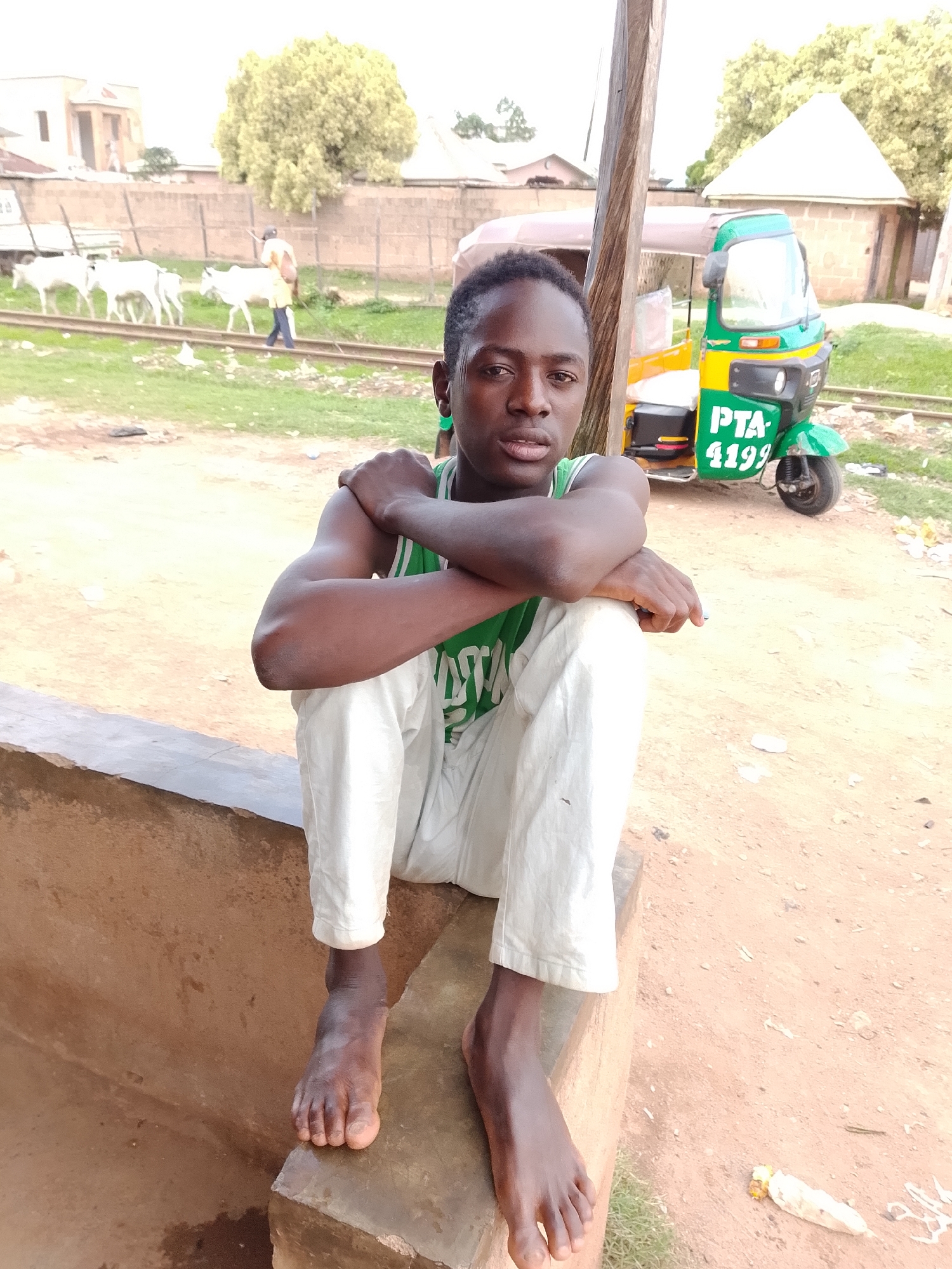 In a dusty, outdoor environment, a young person sits cross-legged on a concrete ledge, facing the camera. They are wearing a green tank top and loose white pants. Their arms are crossed, and their bare feet rest on the ledge. In the background, several white cows are visible near train tracks, and a person in a yellow shirt is walking behind them. A green and yellow auto-rickshaw with the license plate "PTA 4199" is parked to the right of the main subject. Buildings and trees are present in the distance. The ground is a mix of dirt and grass. A white object, possibly a piece of corn husk, lies on the ground in the lower right corner.