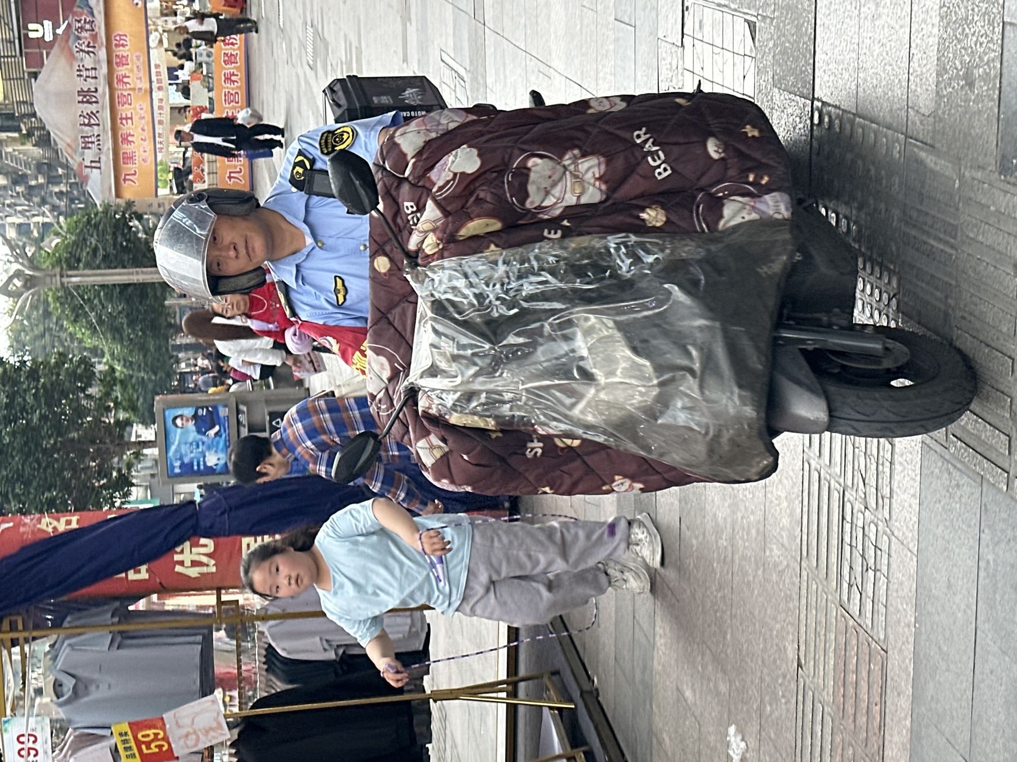The image captures a street scene in what appears to be Jiangjin, China. A man in a police uniform, wearing a clear helmet, sits on a scooter that is partially covered with a patterned blanket featuring bear images. The scooter also has a black box behind the rider. A child is visible in the foreground, jumping rope near a clothing rack with price tags indicating "59" and "99." The backdrop features an advertisement for "Five Black Walnut Nutritional Meal" and "Nine Black Health Nutritional Meal," with Chinese characters and imagery suggesting a bustling urban environment. There are additional people in the background. The ground is paved with concrete tiles.