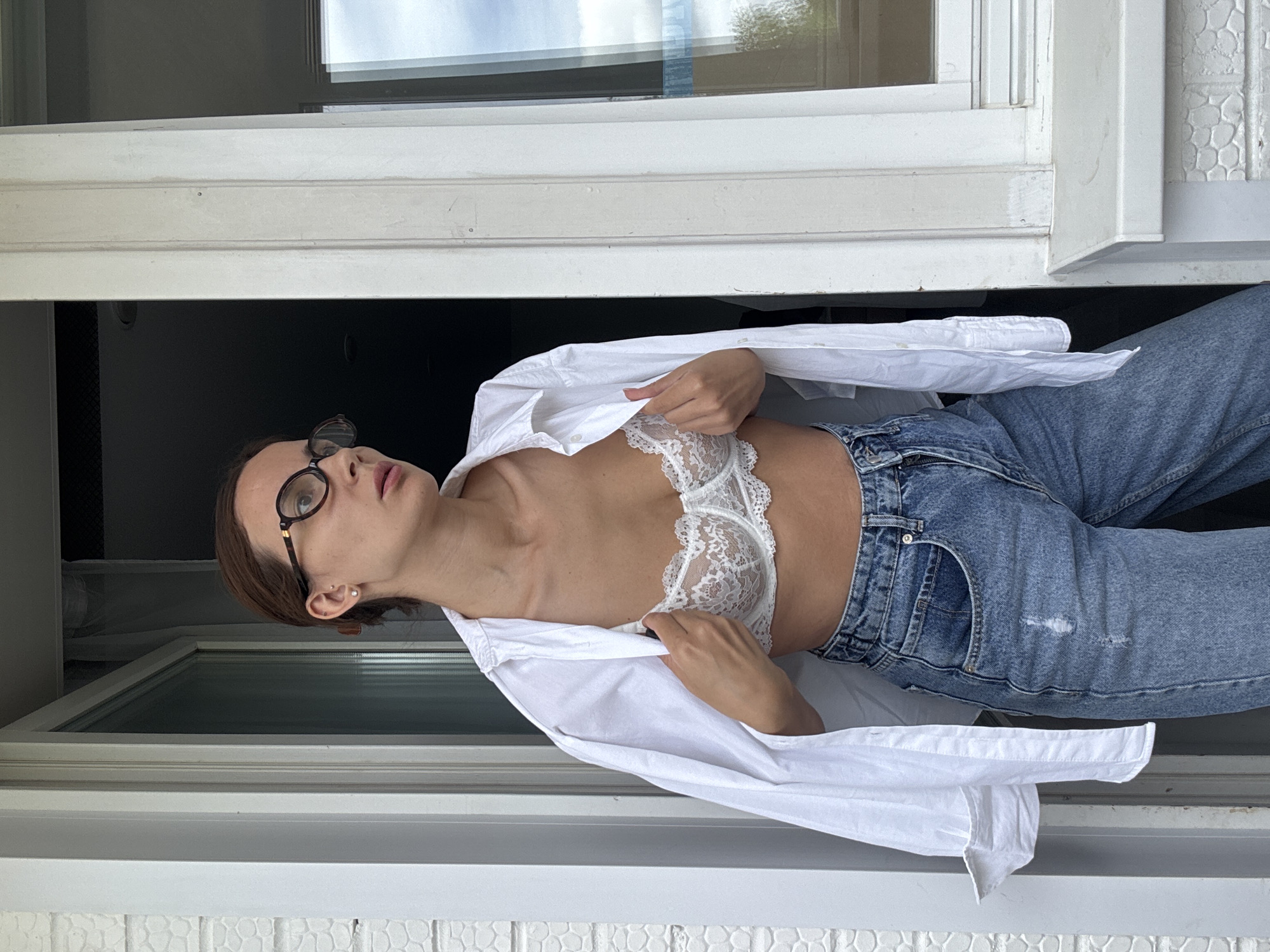 A woman with brown hair pulled back and wearing glasses stands outdoors. She is wearing a white unbuttoned shirt, a white lace bra, and ripped blue jeans. She is holding open the shirt to reveal her bra. The background shows a white building with a window.