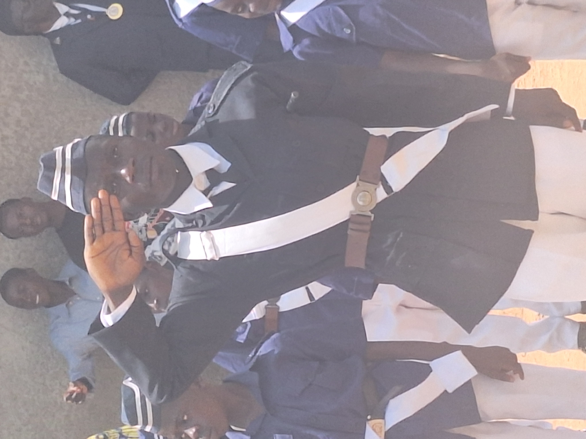 A group of people stand together, appearing to be in a formal or ceremonial setting. Most of the people are dressed in uniforms consisting of a navy blue jacket with white accents, white pants, and a matching cap. One person in the center is raising their right hand, possibly taking an oath or participating in a pledge. A brown belt is worn diagonally across the chest. The location is believed to be Sabon Tasha, Nigeria. The background is slightly blurred.