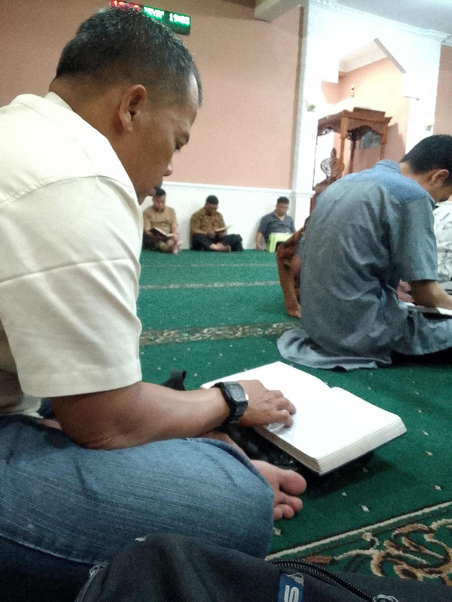 The image depicts a group of men in a room, likely a mosque, based on the green carpet and the presence of what appears to be a prayer niche (mihrab) in the background. The primary subject is a man in the foreground, wearing a light-colored shirt and blue jeans. He is seated on the floor, looking down at an open book. He has a watch on his left wrist. To his left, a black bag with a zipper is visible.
In the middle ground, several other men are seated on the carpet, facing away from the camera or in profile. One man on the left is wearing a brown shirt and holding a book. Another man next to him is also wearing a brown shirt and reading a book. Further back, a man in a dark shirt is seated. On the right side of the image, a man in a blue garment is also seated, partially obscuring the background.
The environment includes a pale pink wall and white trim. A wooden structure, possibly a pulpit or decorative element, is visible in the background. A digital clock displaying red and green lights is mounted high on the wall. The floor is covered with a green carpet with a decorative pattern. The overall scene suggests a religious gathering or study session. The location is identified as Kertajaya, Indonesia.