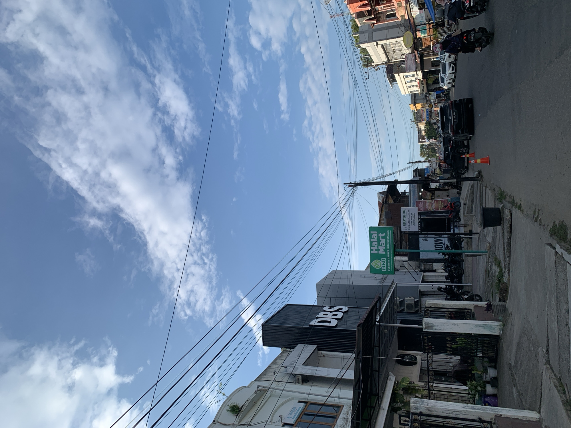 The image depicts a street scene in Lhokseumawe, Indonesia, under a bright, partly cloudy blue sky. Several buildings line the street, some of which appear to be commercial establishments. A prominent sign for "HALAL Mart" is visible, along with other signage for businesses like "DBS" and what appears to be a dental clinic. The street is paved, and there are parked motorcycles and cars. Several power lines crisscross above the street and buildings. On the sidewalk, there are potted plants and a black trash bin. Two individuals are riding motorcycles on the street, one of whom is wearing a headscarf. The overall impression is of an active urban environment during daylight.