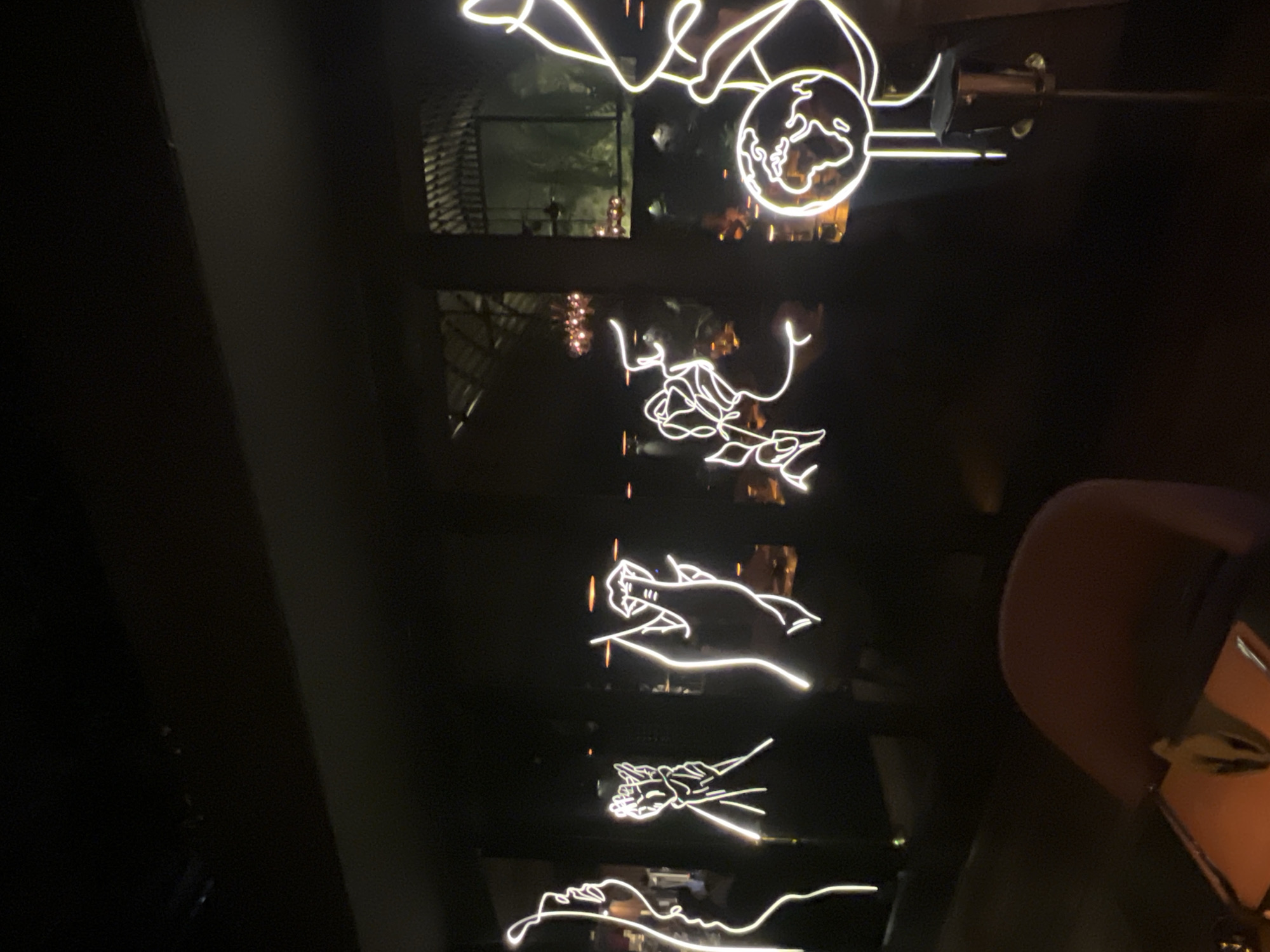 The photograph shows a dimly lit interior space, possibly a restaurant or lounge in North Kuta, Indonesia. Illuminated neon art hangs against a dark background. The neon pieces depict various subjects: a globe, a flower, a hand holding something (perhaps a fish), clasped hands, and a face in profile. Reflections of the outside world are visible through windows behind the neon art. A chair with a reddish-brown back is partially visible in the right side of the image. The overall scene appears to be captured at night.