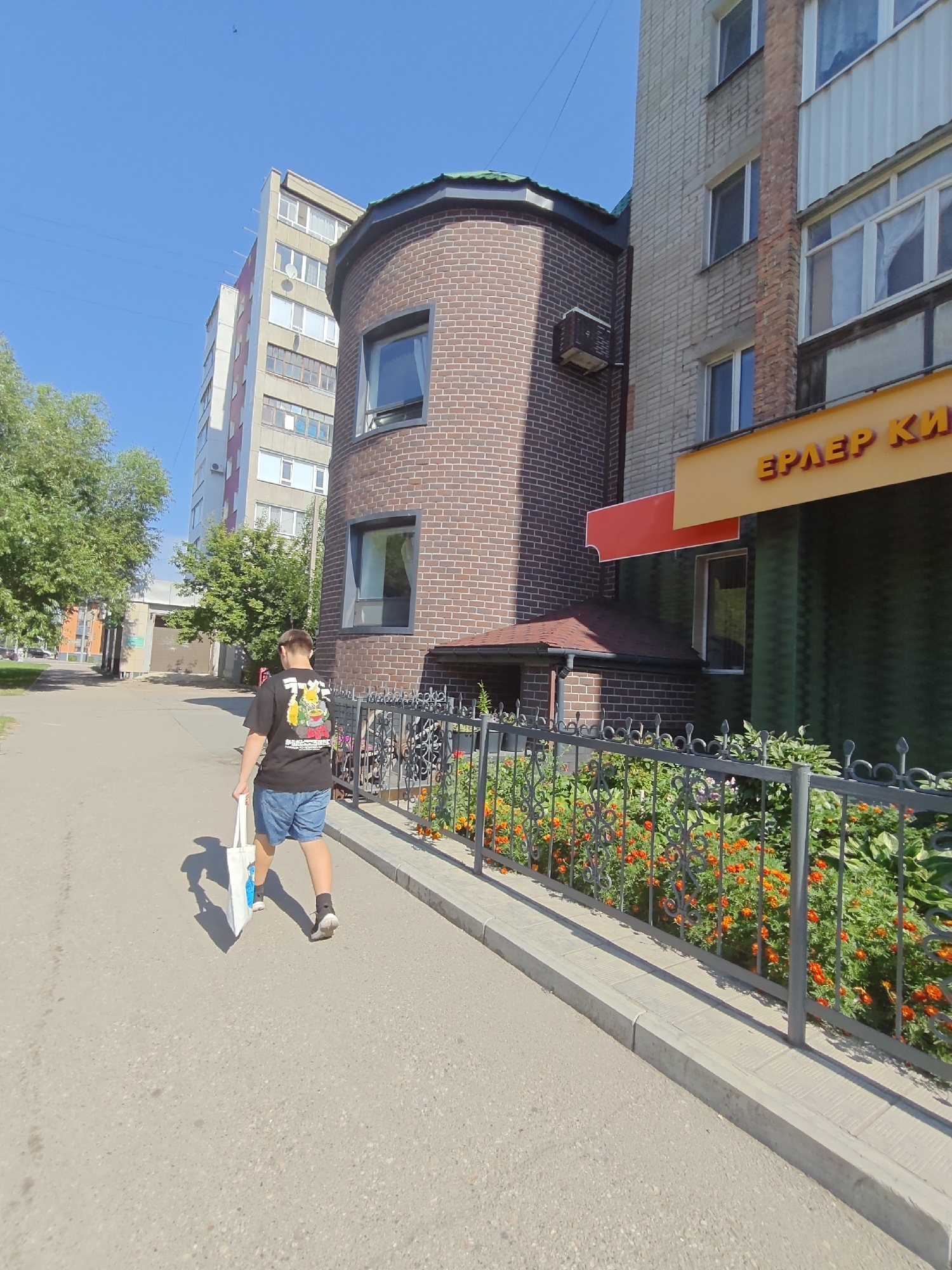 A young person walks away from the camera on a paved sidewalk in front of a multi-story building. The person is wearing a black t-shirt with a colorful graphic on the back and blue denim shorts. They are carrying a white tote bag in their right hand. The sidewalk is bordered by a decorative metal fence with a flower bed behind it. The flower bed contains numerous small, bright orange flowers and green foliage. The building behind the fence is made of dark red brick with a rounded section. Above the brick section, a light-colored multi-story building with many windows is visible. A yellow sign with red Cyrillic lettering is mounted on the side of the building. The sky is clear and blue. This scene is located on a street in the city of Petropavl, Kazakhstan.