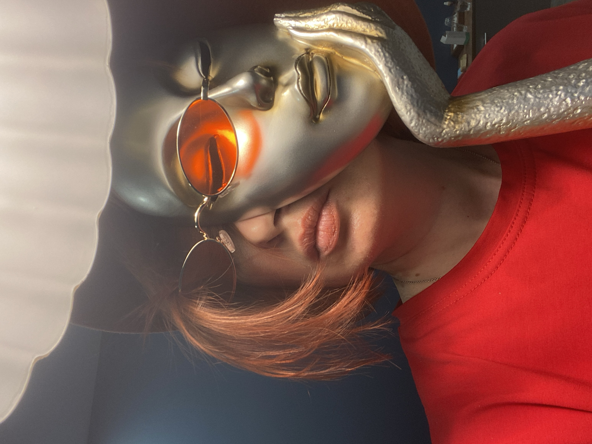 A close-up, rotated image shows a person in a red shirt partially obscured by a metallic mask wearing round, reddish-tinted sunglasses. The person's lower face is visible, showing their lips and part of their nose, with reddish-brown hair falling across their forehead. A white, scalloped edge is visible on the left side of the frame. The background is dark, appearing to be a wall. There is a shiny, metallic arm or extension of the mask draped across the top. There is no information in the image to indicate location, nor is it possible to determine the specific city or country from the image alone.