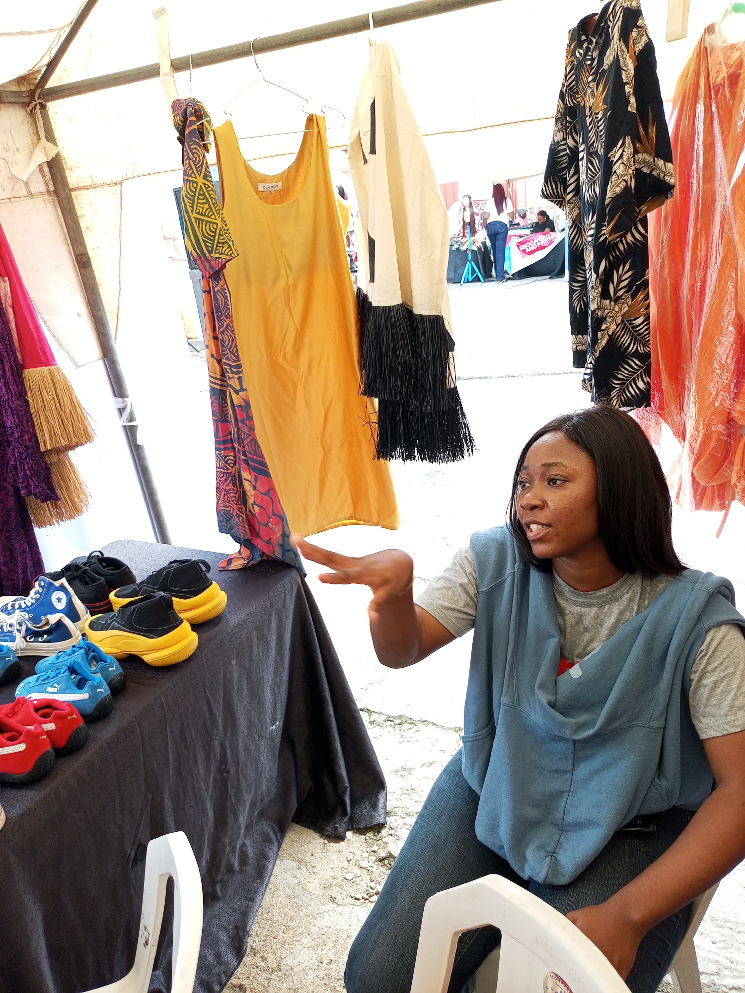 Ikeja, Nigeria. A woman sits beside a table covered with a black cloth upon which several pairs of shoes are displayed. Clothes are hanging on a rack behind her. The woman is dark-skinned and wears jeans, a t-shirt, and a hooded sweatshirt. Her right hand is extended.