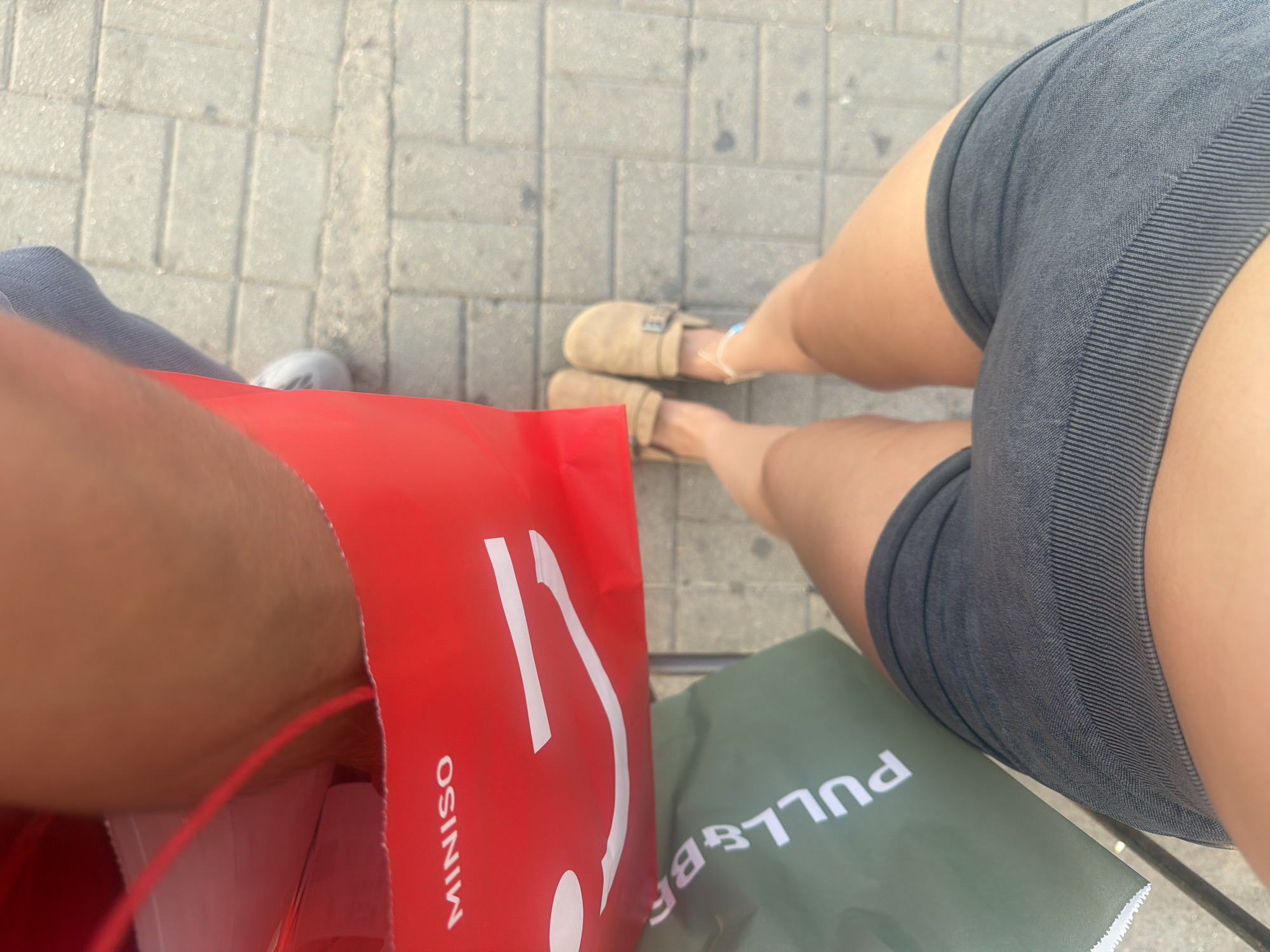 A first-person perspective shot shows a person's legs and feet, along with shopping bags, on a paved surface. The person is wearing dark gray shorts and tan clogs. In front of the person are two shopping bags: one red bag with "MINISO" written on it and a smiley face, and a green bag with "Pull&Bear" visible. The paved surface is made of light gray rectangular stones. In the background, a person's arm and shoulder, wearing a blue garment, are visible, suggesting the person is sitting. The lighting indicates it is daytime, with harsh shadows. The scene appears to be outdoors, possibly in a pedestrian area or street.
