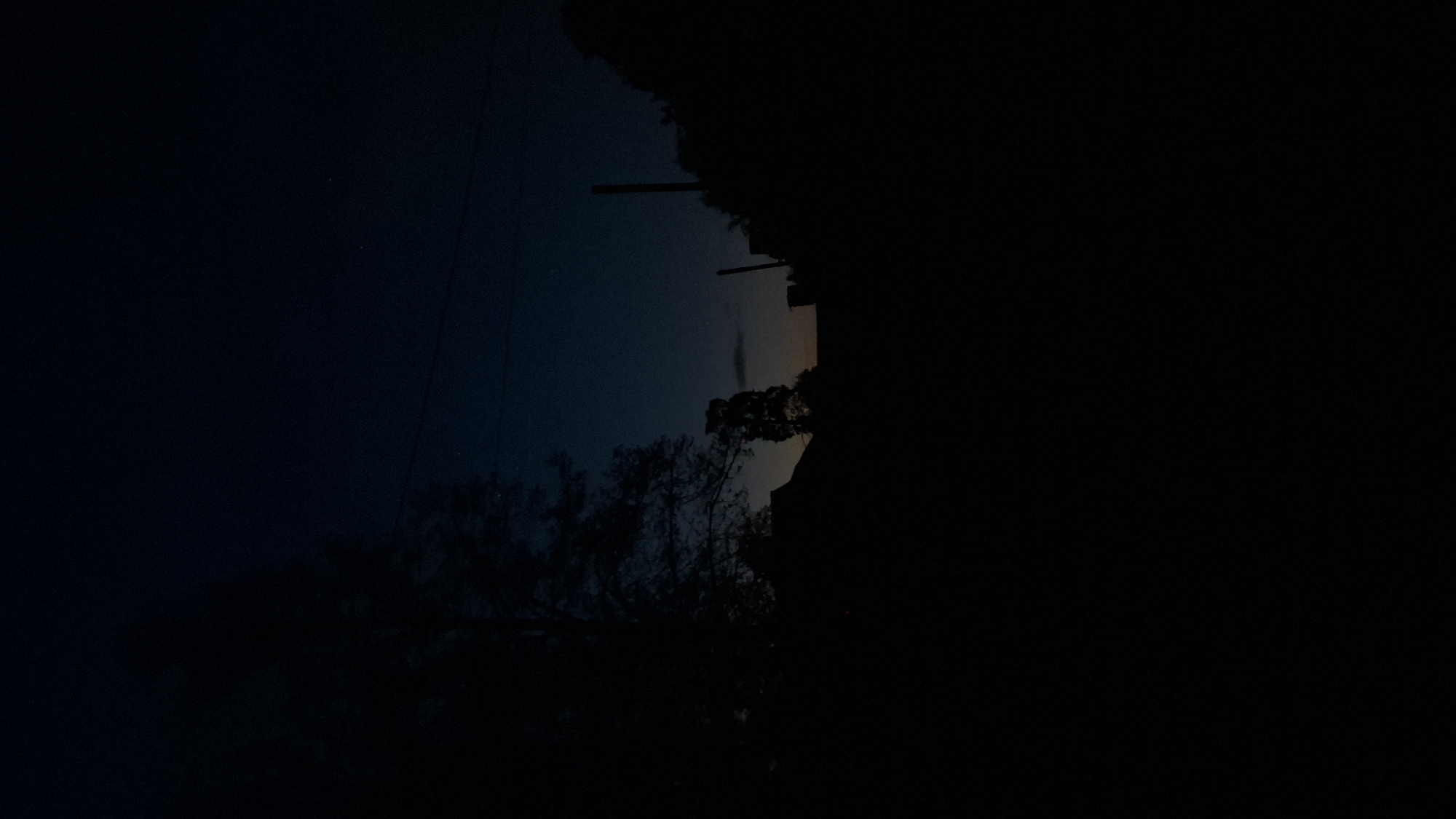 The image depicts a night scene in Шешурга, Russia. The left portion of the image shows a dark blue sky with faint stars and silhouetted trees and power lines. The right portion is predominantly black, obscuring details, although a building's silhouette and a tree are discernible. The image is rotated to the left.