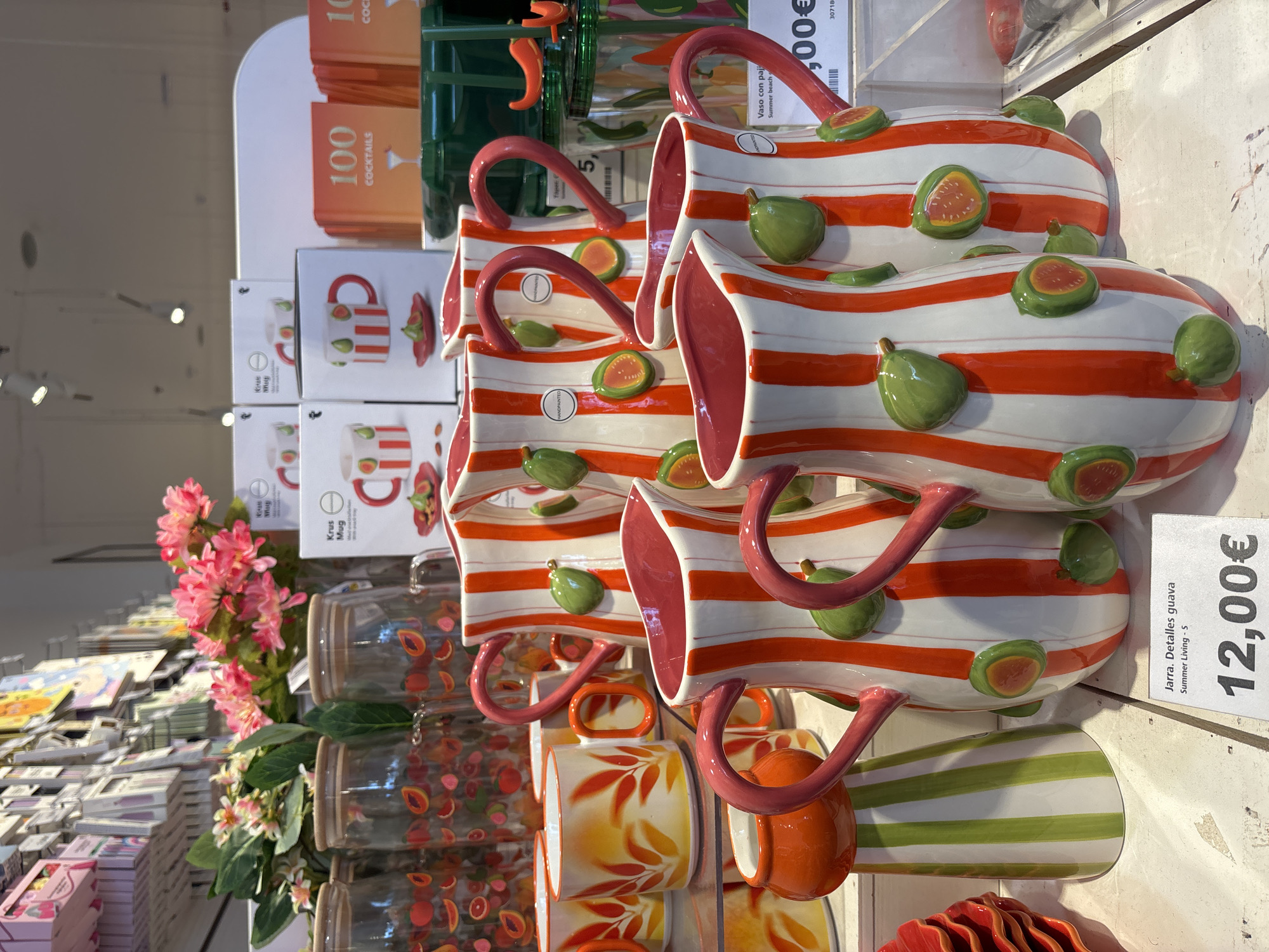 The image shows a retail display of ceramic pitchers and mugs with a fruit motif, specifically guavas and possibly citrus. The pitchers are tall and cylindrical with red and white vertical stripes, and are decorated with raised green guava shapes. They have bright pink handles. Several mugs are also visible, some with similar red and white stripes, and others with a yellow and orange leaf pattern. In the background, there are glass jars with a similar fruit pattern and stacks of books titled "100 Cocktails". A price tag for one of the pitchers reads "Jarra. Detalles guava Summer Living - S 12,00 €". The display appears to be in a well-lit store, possibly a home goods or gift shop. The overall impression is one of vibrant, summer-themed tableware. The provided context suggests this scene is located in Orihuela, Spain.