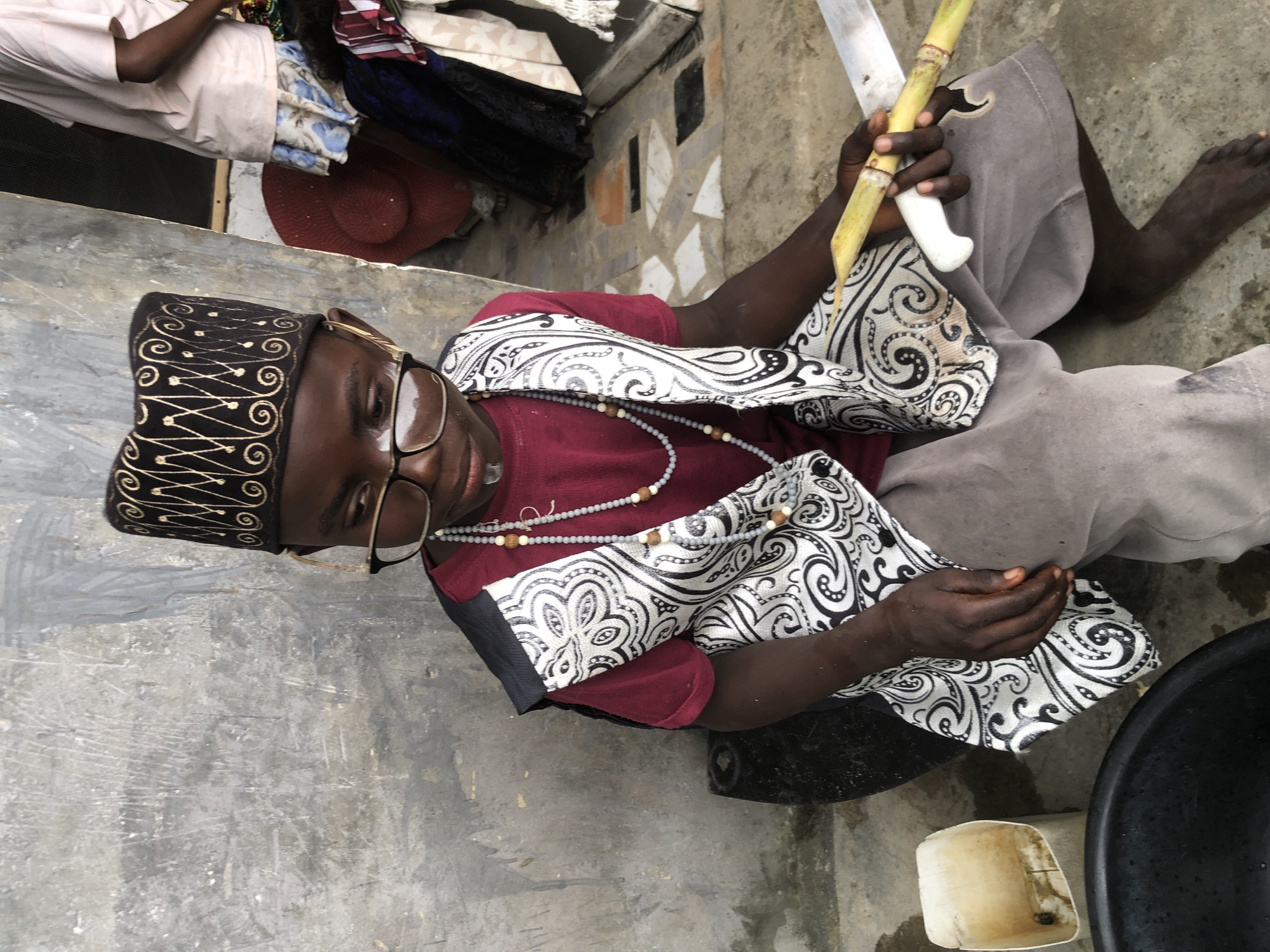 A young boy with dark skin, wearing a maroon t-shirt, a decorative vest with a black and white swirling pattern, and a black cap with gold embroidery, sits on the ground in what appears to be an outdoor, urban setting in Ado, Nigeria. He has on glasses and is holding a piece of sugarcane in his left hand, while his right hand holds a large knife, positioned to cut the sugarcane. To his left is a white plastic container. In the background, a wall with a rough, grey texture is visible, and further back, another person in a pink t-shirt and a patterned skirt stands near some fabrics and a red hat. The ground is concrete with a few scattered stones. The overall scene suggests a moment of everyday life, possibly involving the preparation of food or a craft.